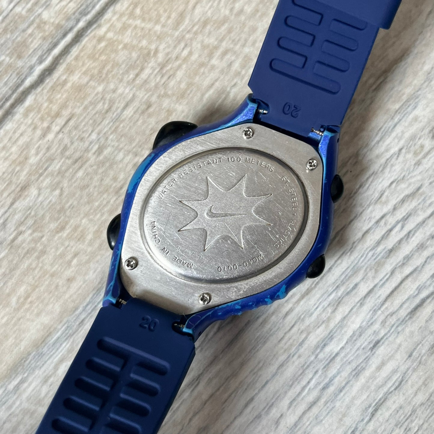 Nike Triax 120 WG40-0010 Digital Watch features a blue case with a black Nike Swoosh above the display and a black control button below it. The watch has a black digital screen with light-colored numerals and is fitted with navy-blue replacement straps that are not original Nike straps. Constructed from stainless steel and plastic materials, it offers 100-meter water resistance, 120-lap memory, and multiple timing and training modes.