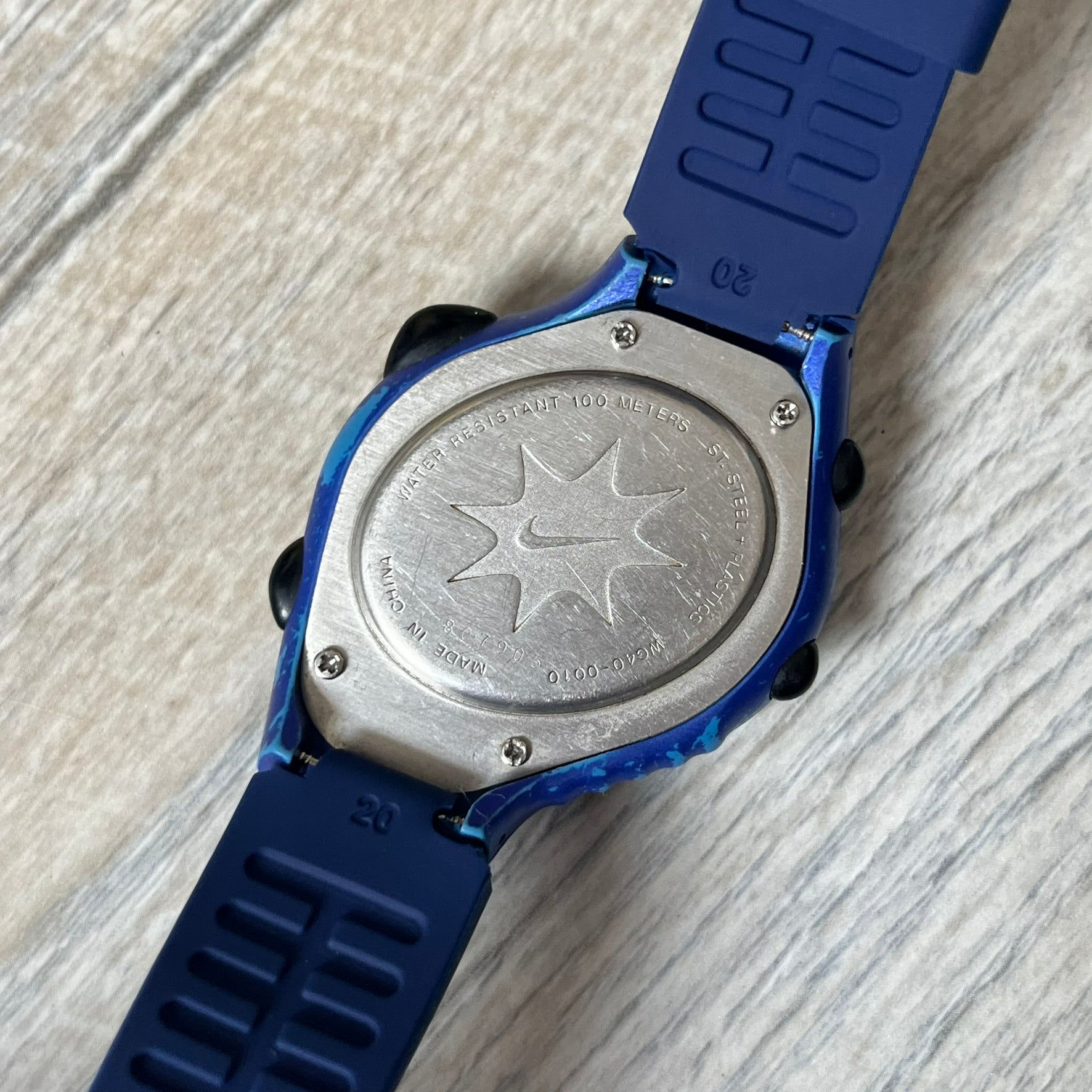 Nike Triax 120 WG40-0010 Digital Watch features a blue case with a black Nike Swoosh above the display and a black control button below it. The watch has a black digital screen with light-colored numerals and is fitted with navy-blue replacement straps that are not original Nike straps. Constructed from stainless steel and plastic materials, it offers 100-meter water resistance, 120-lap memory, and multiple timing and training modes.