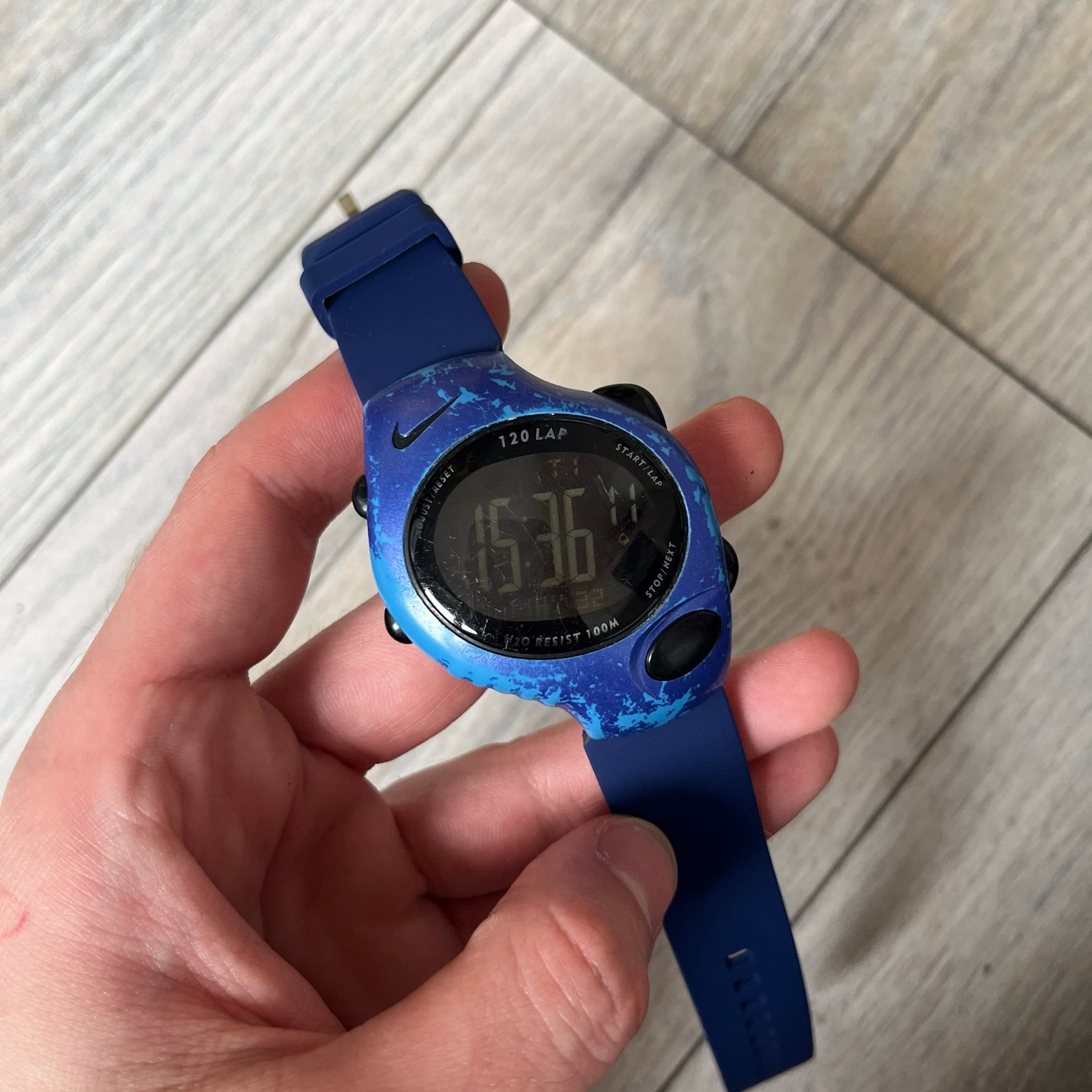 Nike Triax 120 WG40-0010 Digital Watch features a blue case with a black Nike Swoosh above the display and a black control button below it. The watch has a black digital screen with light-colored numerals and is fitted with navy-blue replacement straps that are not original Nike straps. Constructed from stainless steel and plastic materials, it offers 100-meter water resistance, 120-lap memory, and multiple timing and training modes.