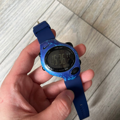 Nike Triax 120 WG40-0010 Digital Watch features a blue case with a black Nike Swoosh above the display and a black control button below it. The watch has a black digital screen with light-colored numerals and is fitted with navy-blue replacement straps that are not original Nike straps. Constructed from stainless steel and plastic materials, it offers 100-meter water resistance, 120-lap memory, and multiple timing and training modes.