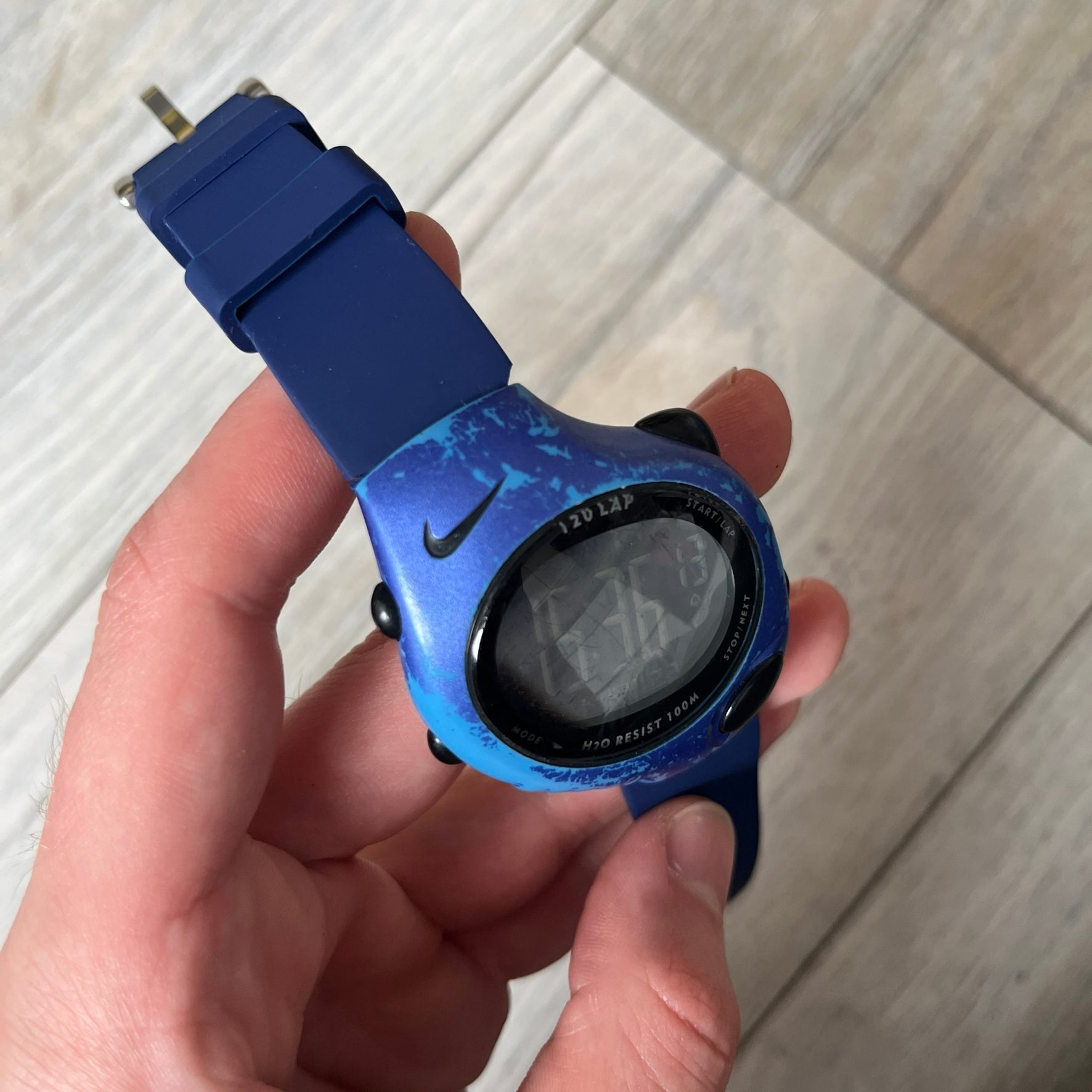 Nike Triax 120 WG40-0010 Digital Watch features a blue case with a black Nike Swoosh above the display and a black control button below it. The watch has a black digital screen with light-colored numerals and is fitted with navy-blue replacement straps that are not original Nike straps. Constructed from stainless steel and plastic materials, it offers 100-meter water resistance, 120-lap memory, and multiple timing and training modes.