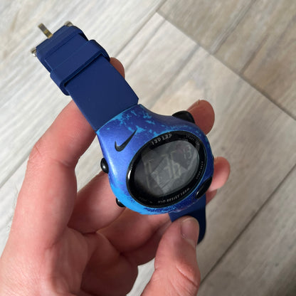 Nike Triax 120 WG40-0010 Digital Watch features a blue case with a black Nike Swoosh above the display and a black control button below it. The watch has a black digital screen with light-colored numerals and is fitted with navy-blue replacement straps that are not original Nike straps. Constructed from stainless steel and plastic materials, it offers 100-meter water resistance, 120-lap memory, and multiple timing and training modes.