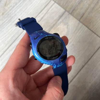 Nike Triax 120 WG40-0010 Digital Watch features a blue case with a black Nike Swoosh above the display and a black control button below it. The watch has a black digital screen with light-colored numerals and is fitted with navy-blue replacement straps that are not original Nike straps. Constructed from stainless steel and plastic materials, it offers 100-meter water resistance, 120-lap memory, and multiple timing and training modes.