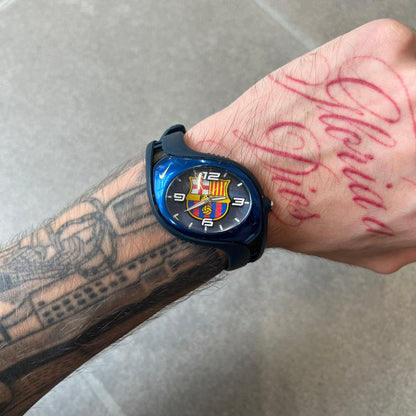 Nike Triax 2005 FC Barcelona WK0008 Analog Watch is a vintage watch featuring a navy rubber strap, metal case and dial. The dial displays a large full-color FC Barcelona crest with white numerals, while a yellow Nike Swoosh is positioned at the top of the case. The watch is water resistant up to 50 meters and is constructed from stainless steel and plastic materials.