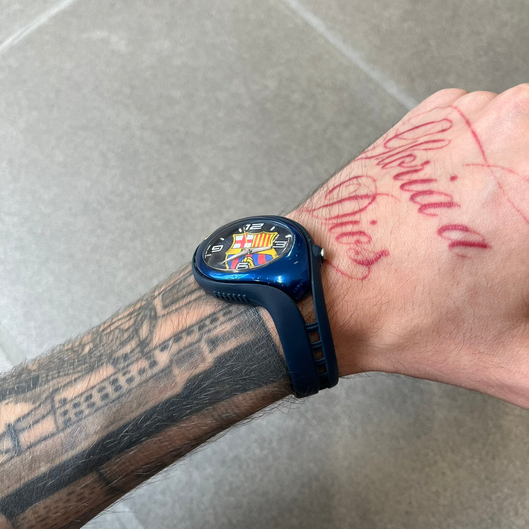 Nike Triax 2005 FC Barcelona WK0008 Analog Watch is a vintage watch featuring a navy rubber strap, metal case and dial. The dial displays a large full-color FC Barcelona crest with white numerals, while a yellow Nike Swoosh is positioned at the top of the case. The watch is water resistant up to 50 meters and is constructed from stainless steel and plastic materials.