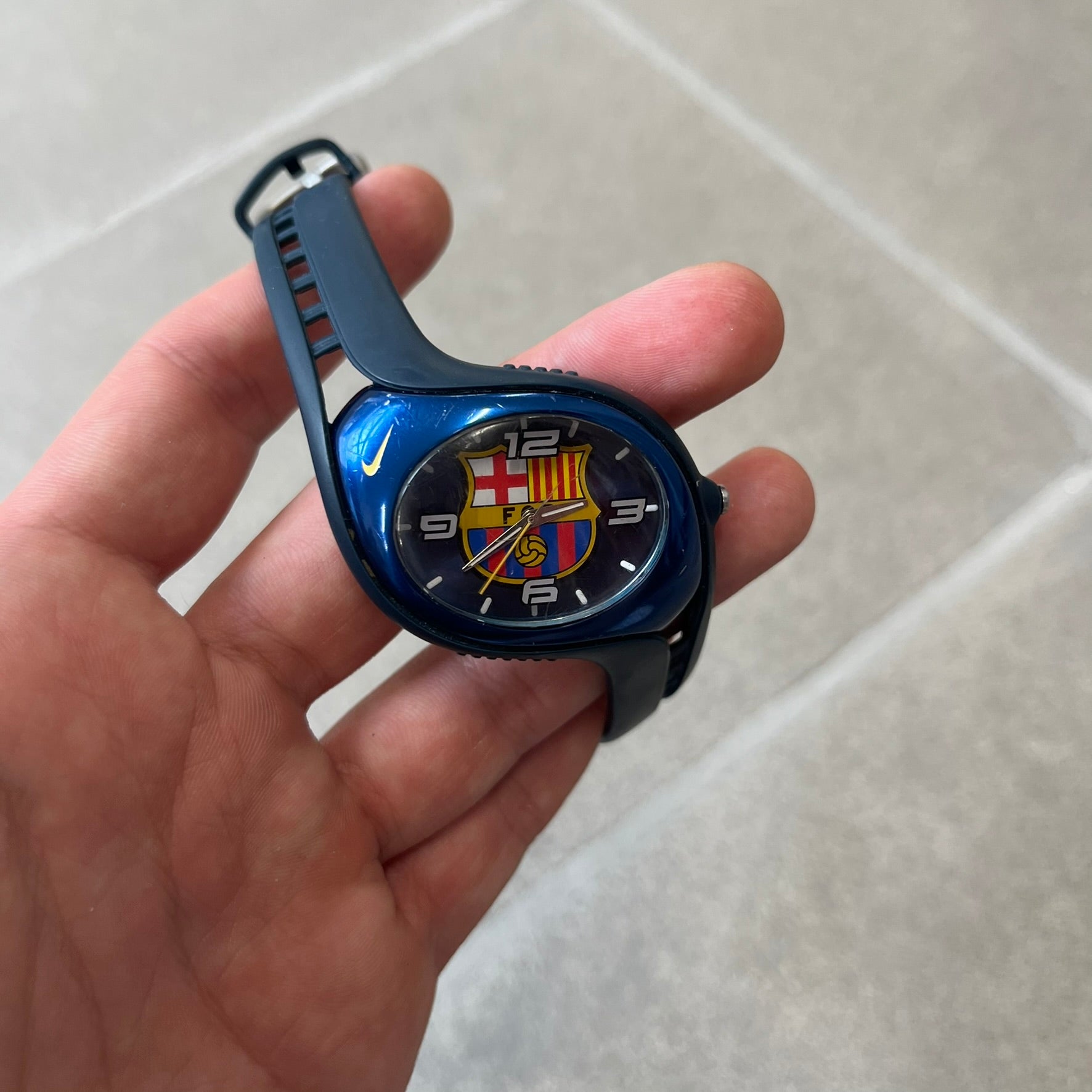 Nike Triax 2005 FC Barcelona WK0008 Analog Watch is a vintage watch featuring a navy rubber strap, metal case and dial. The dial displays a large full-color FC Barcelona crest with white numerals, while a yellow Nike Swoosh is positioned at the top of the case. The watch is water resistant up to 50 meters and is constructed from stainless steel and plastic materials.