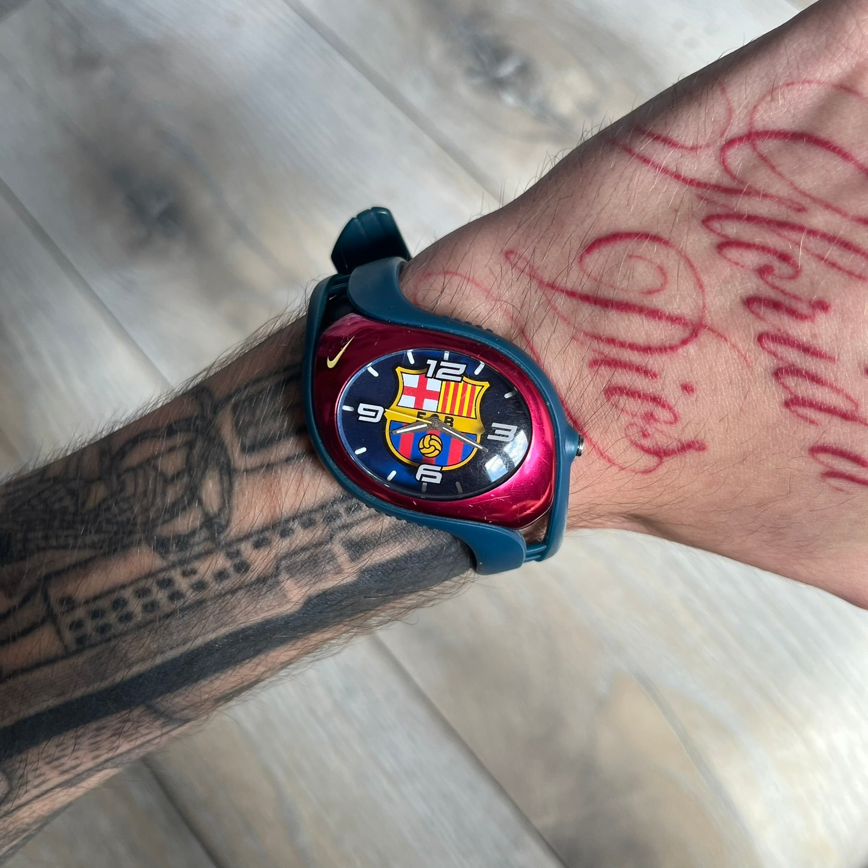 Nike Triax 2005 FC Barcelona WK0008 Analog Watch is a vintage watch featuring a navy rubber strap, red plastic case, and navy dial. The dial displays a large full-color FC Barcelona crest with white numerals, while a yellow Nike Swoosh is positioned at the top of the case. The watch is water resistant up to 50 meters and is constructed from stainless steel and plastic materials.