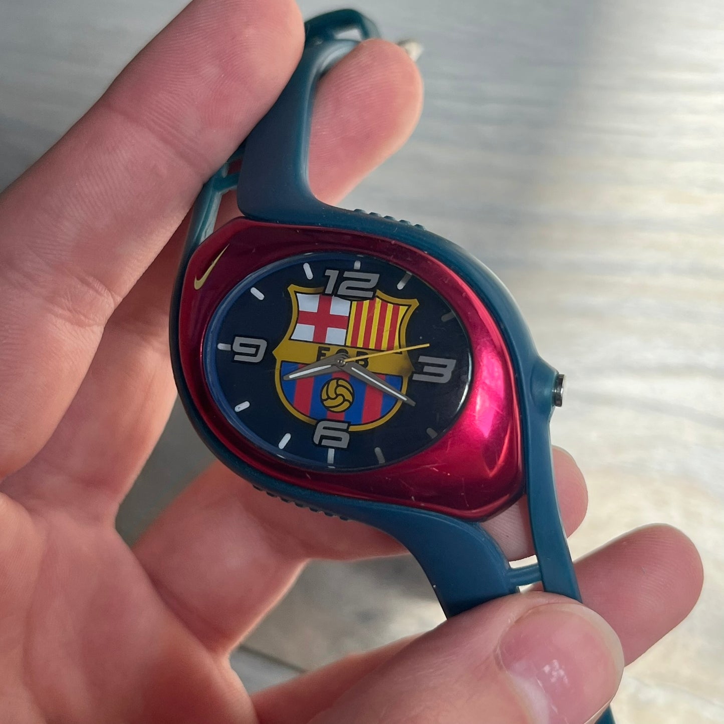Nike Triax 2005 FC Barcelona WK0008 Analog Watch is a vintage watch featuring a navy rubber strap, red plastic case, and navy dial. The dial displays a large full-color FC Barcelona crest with white numerals, while a yellow Nike Swoosh is positioned at the top of the case. The watch is water resistant up to 50 meters and is constructed from stainless steel and plastic materials.