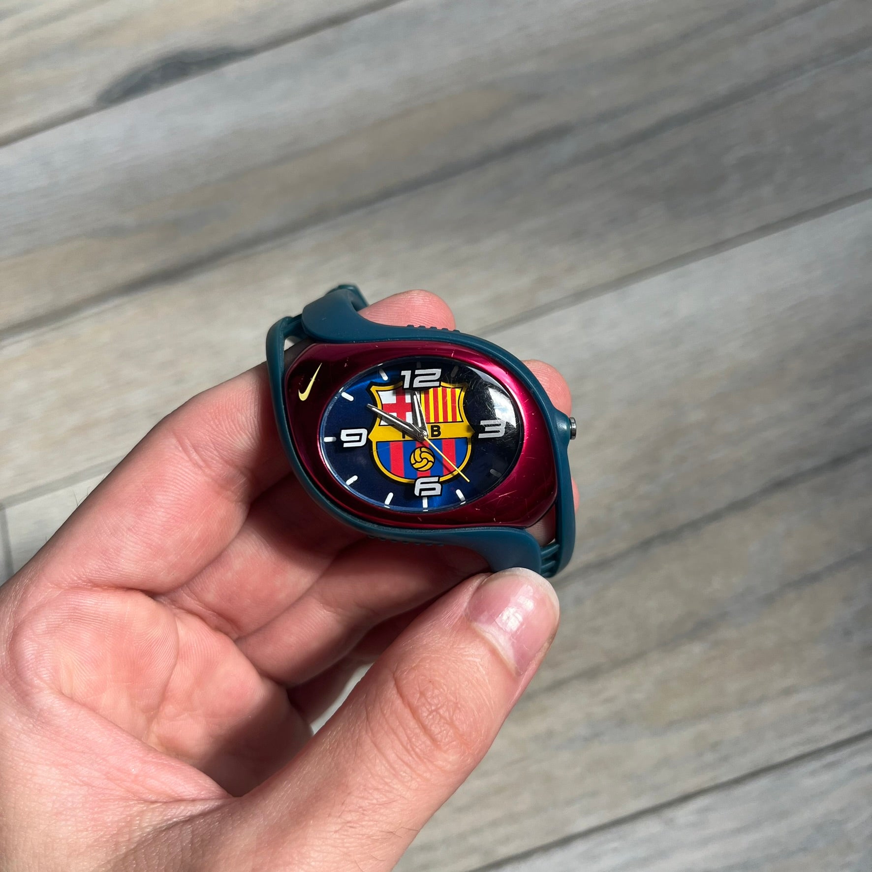 Nike Triax 2005 FC Barcelona WK0008 Analog Watch is a vintage watch featuring a navy rubber strap, red plastic case, and navy dial. The dial displays a large full-color FC Barcelona crest with white numerals, while a yellow Nike Swoosh is positioned at the top of the case. The watch is water resistant up to 50 meters and is constructed from stainless steel and plastic materials.