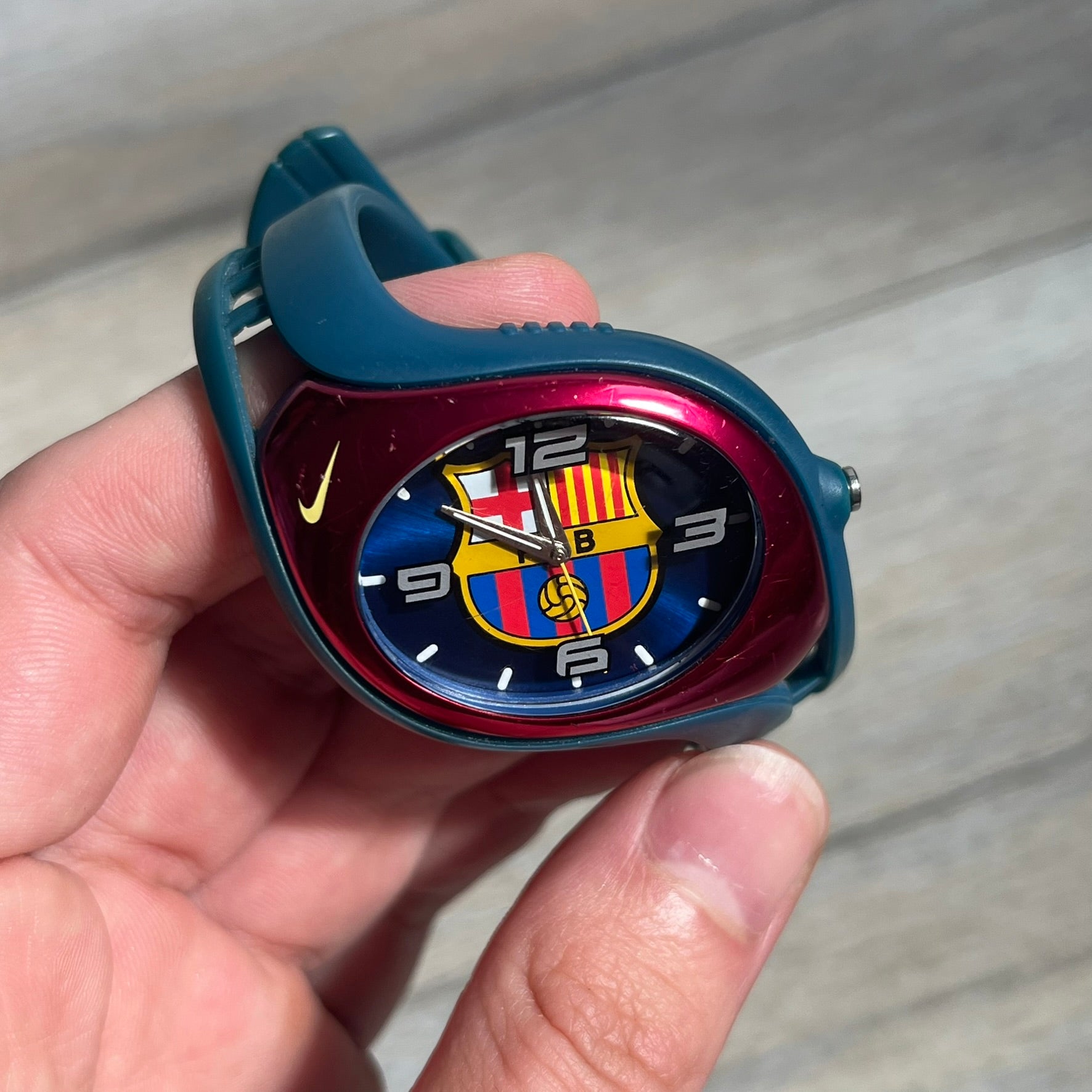 Nike Triax 2005 FC Barcelona WK0008 Analog Watch is a vintage watch featuring a navy rubber strap, red plastic case, and navy dial. The dial displays a large full-color FC Barcelona crest with white numerals, while a yellow Nike Swoosh is positioned at the top of the case. The watch is water resistant up to 50 meters and is constructed from stainless steel and plastic materials.