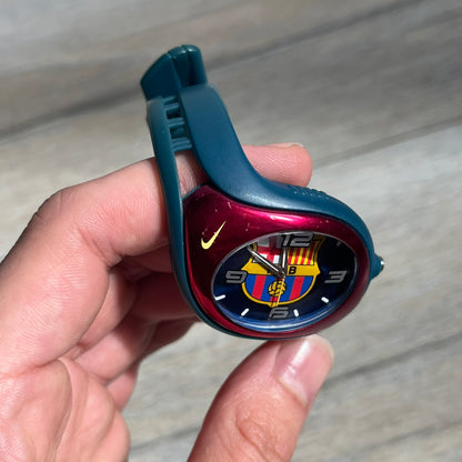 Nike Triax 2005 FC Barcelona WK0008 Analog Watch is a vintage watch featuring a navy rubber strap, red plastic case, and navy dial. The dial displays a large full-color FC Barcelona crest with white numerals, while a yellow Nike Swoosh is positioned at the top of the case. The watch is water resistant up to 50 meters and is constructed from stainless steel and plastic materials.