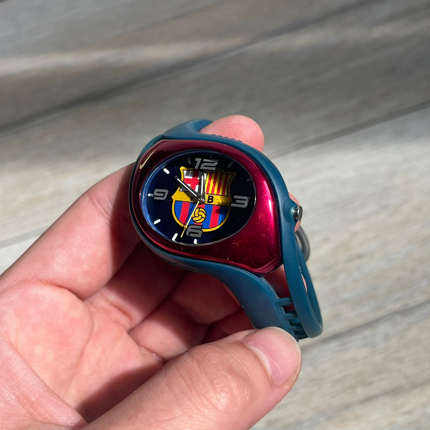 Nike Triax 2005 FC Barcelona WK0008 Analog Watch is a vintage watch featuring a navy rubber strap, red plastic case, and navy dial. The dial displays a large full-color FC Barcelona crest with white numerals, while a yellow Nike Swoosh is positioned at the top of the case. The watch is water resistant up to 50 meters and is constructed from stainless steel and plastic materials.