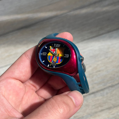 Nike Triax 2005 FC Barcelona WK0008 Analog Watch is a vintage watch featuring a navy rubber strap, red plastic case, and navy dial. The dial displays a large full-color FC Barcelona crest with white numerals, while a yellow Nike Swoosh is positioned at the top of the case. The watch is water resistant up to 50 meters and is constructed from stainless steel and plastic materials.