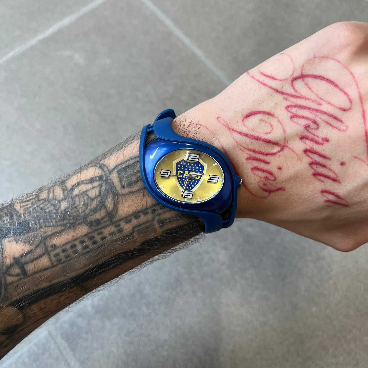 Nike Triax Blaze Boca Juniors WD0055 Analog Watch is a vintage watch featuring a blue rubber strap, metal case, and yellow dial. The dial displays a large full-color Boca Juniors crest with white main numerals, while a yellow Nike Swoosh is positioned at the top of the case. The watch is water resistant up to 50 meters and is constructed from stainless steel and plastic materials.
