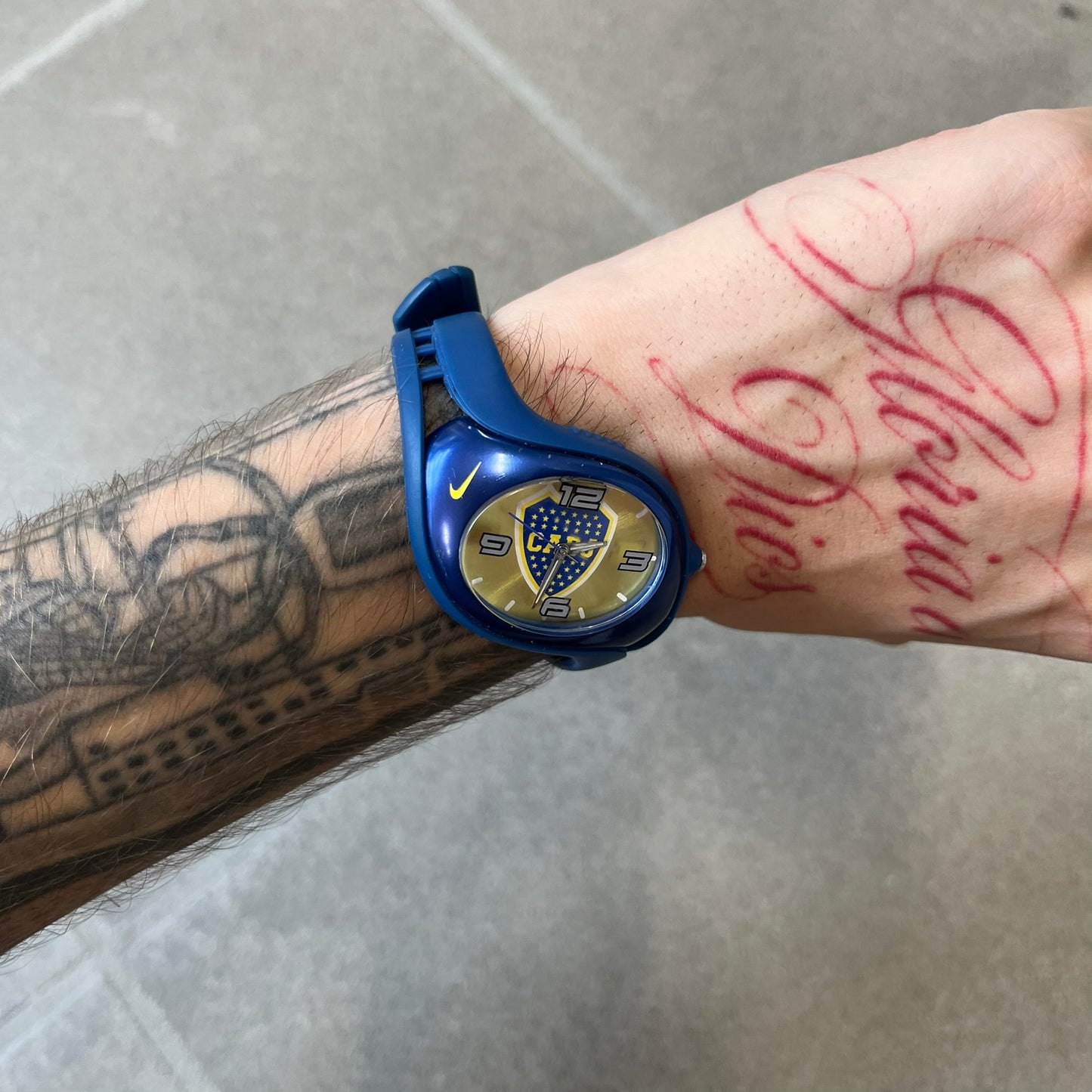 Nike Triax Blaze Boca Juniors WD0055 Analog Watch is a vintage watch featuring a blue rubber strap, metal case, and yellow dial. The dial displays a large full-color Boca Juniors crest with white main numerals, while a yellow Nike Swoosh is positioned at the top of the case. The watch is water resistant up to 50 meters and is constructed from stainless steel and plastic materials.