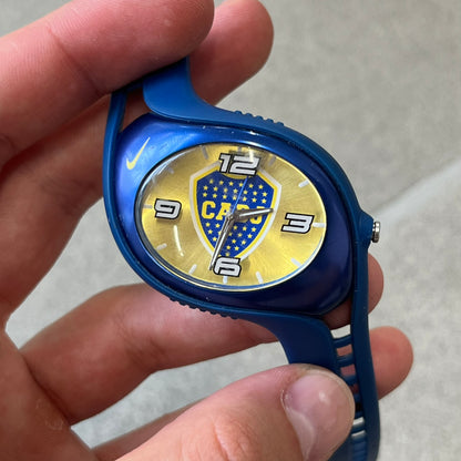 Nike Triax Blaze Boca Juniors WD0055 Analog Watch is a vintage watch featuring a blue rubber strap, metal case, and yellow dial. The dial displays a large full-color Boca Juniors crest with white main numerals, while a yellow Nike Swoosh is positioned at the top of the case. The watch is water resistant up to 50 meters and is constructed from stainless steel and plastic materials.