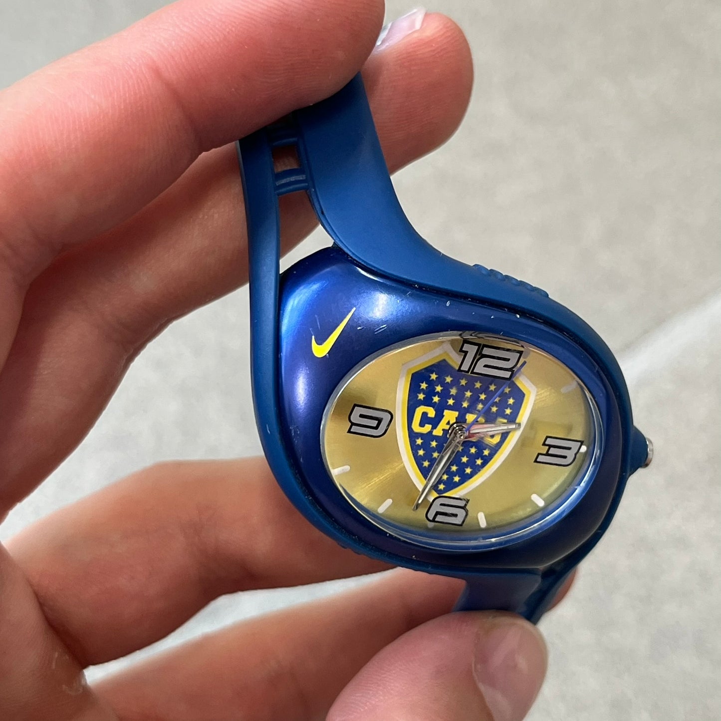 Nike Triax Blaze Boca Juniors WD0055 Analog Watch is a vintage watch featuring a blue rubber strap, metal case, and yellow dial. The dial displays a large full-color Boca Juniors crest with white main numerals, while a yellow Nike Swoosh is positioned at the top of the case. The watch is water resistant up to 50 meters and is constructed from stainless steel and plastic materials.