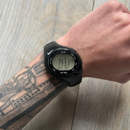 The Nike Triax Bowerman WR0124 Black Digital Watch features black rubber straps and a black plastic case with a metal caseback engraved with the Bowerman logo. It includes a digital display with multiple modes, a 10-lap memory function, H2O 100 water resistance, and a built-in backlight. The watch has a single front button and a white Nike logo on the case.