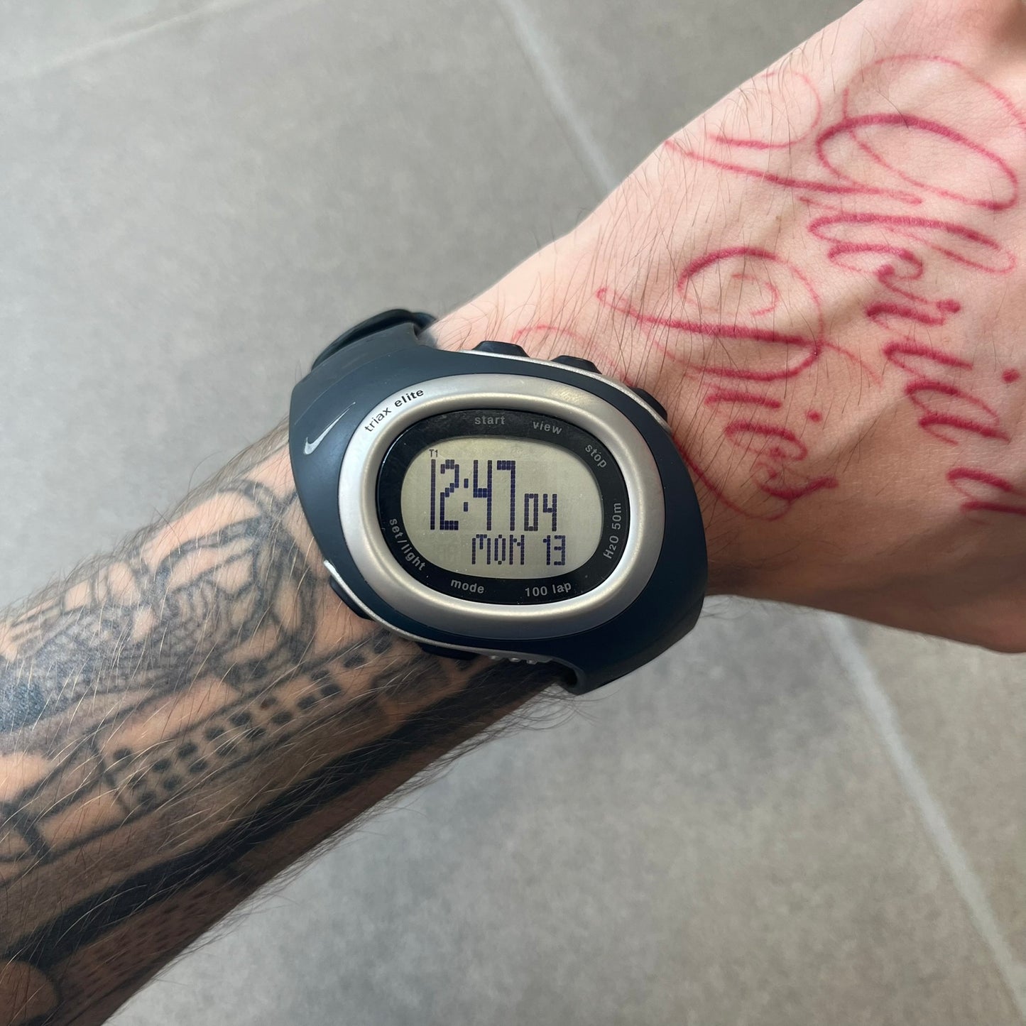 Nike Triax Elite SM0019 Digital Watch features a large asymmetrical silver-tone case with navy rubber case and straps integrated into the case. The top of the case is detailed with a Nike Swoosh, while the front includes Triax Elite branding. The watch has five side buttons, a digital display with multiple functions, and 100-lap memory. It comes with an untested Nike heart rate monitor (HRM) device and an original Nike watch display stand.