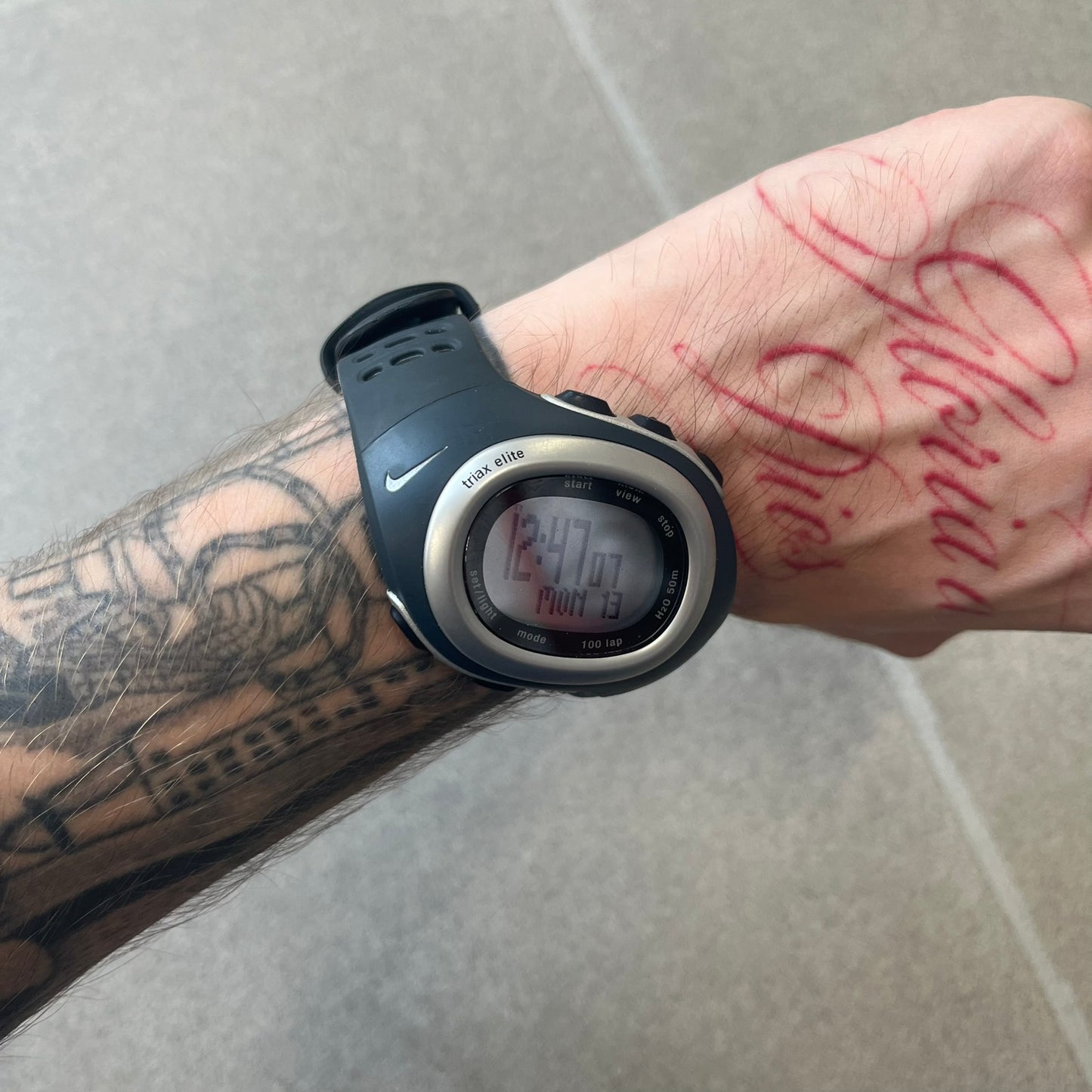 Nike Triax Elite SM0019 Digital Watch features a large asymmetrical silver-tone case with navy rubber case and straps integrated into the case. The top of the case is detailed with a Nike Swoosh, while the front includes Triax Elite branding. The watch has five side buttons, a digital display with multiple functions, and 100-lap memory. It comes with an untested Nike heart rate monitor (HRM) device and an original Nike watch display stand.