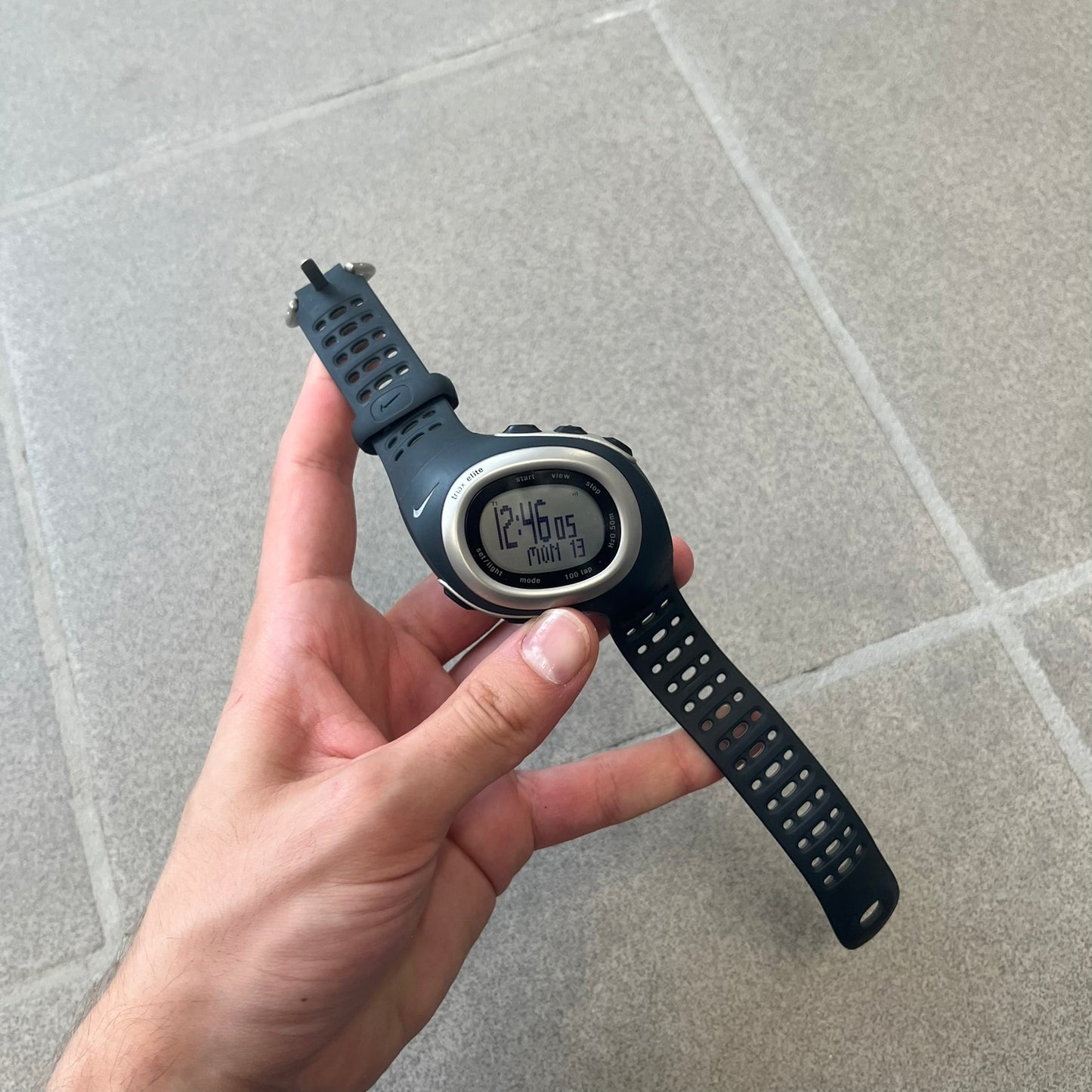 Nike Triax Elite SM0019 Digital Watch features a large asymmetrical silver-tone case with navy rubber case and straps integrated into the case. The top of the case is detailed with a Nike Swoosh, while the front includes Triax Elite branding. The watch has five side buttons, a digital display with multiple functions, and 100-lap memory. It comes with an untested Nike heart rate monitor (HRM) device and an original Nike watch display stand.
