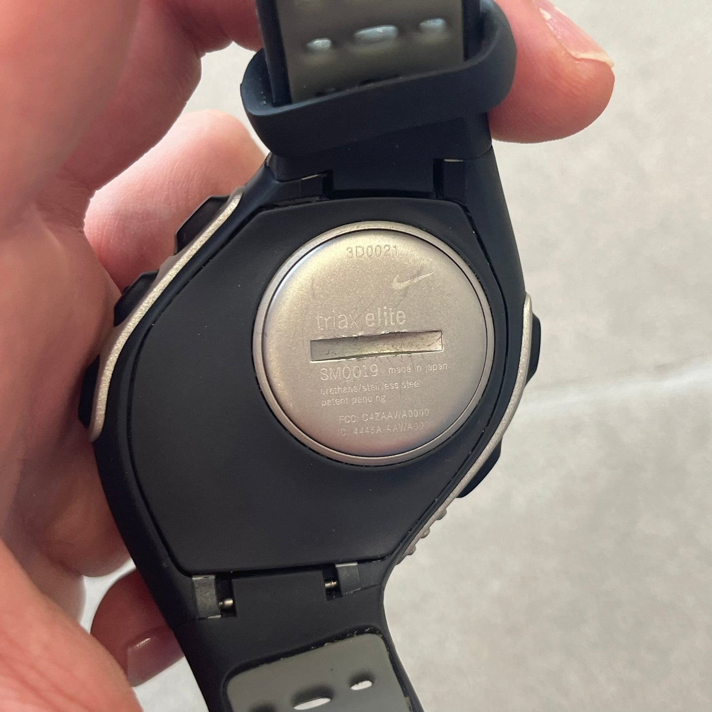 Nike Triax Elite SM0019 Digital Watch features a large asymmetrical silver-tone case with navy rubber case and straps integrated into the case. The top of the case is detailed with a Nike Swoosh, while the front includes Triax Elite branding. The watch has five side buttons, a digital display with multiple functions, and 100-lap memory. It comes with an untested Nike heart rate monitor (HRM) device and an original Nike watch display stand.