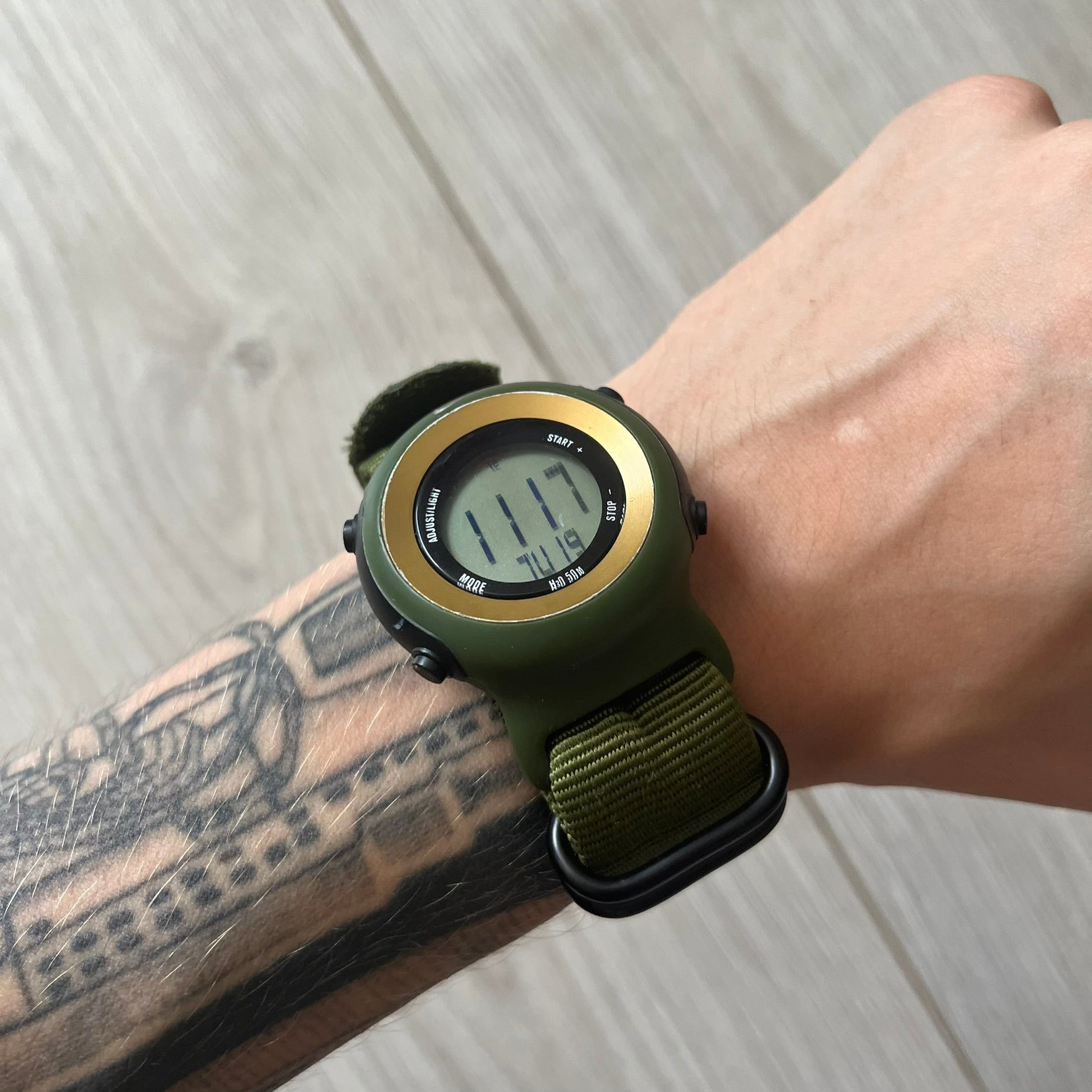 Green digital watch on a wrist with a tattooed arm against a light wooden background