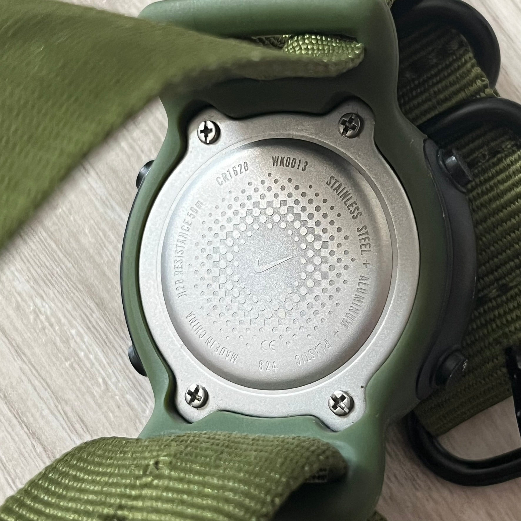 Close-up of a green and silver watch with a textured metal back on a light wooden surface.