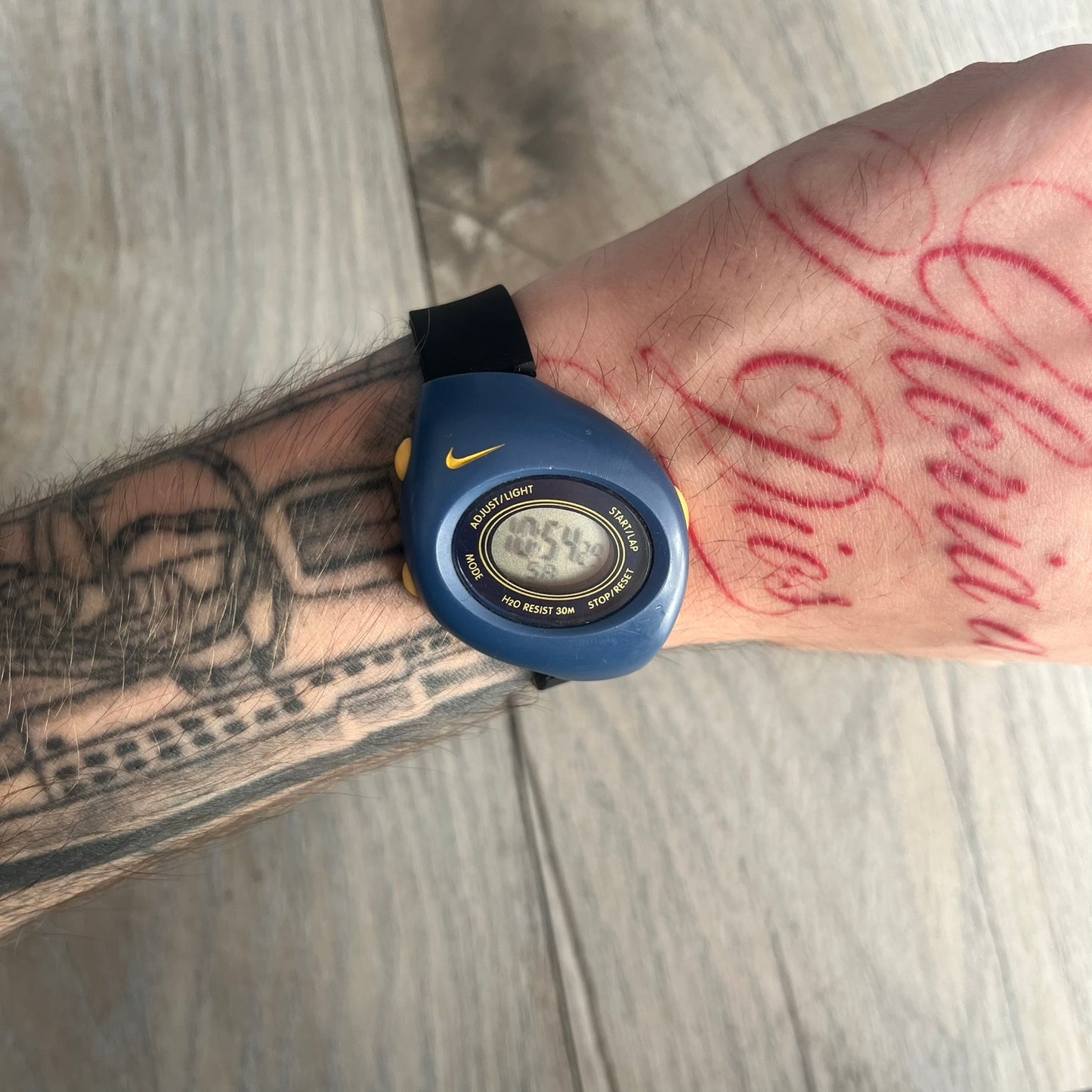Nike Triax Junior 2000s Blue Digital Watch is a digital timepiece featuring a blue case with a yellow Nike Swoosh on the bezel. The watch includes a backlight with the Bowerman Nike logo, yellow side buttons, and a navy ring around the display with yellow numerals and markings. It has a stainless steel and plastic construction with 30m water resistance. The original strap has been replaced with a black non-Nike strap.
