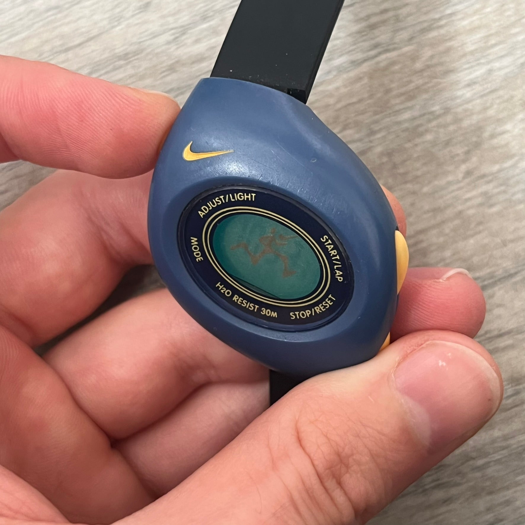 Nike Triax Junior 2000s Blue Digital Watch is a digital timepiece featuring a blue case with a yellow Nike Swoosh on the bezel. The watch includes a backlight with the Bowerman Nike logo, yellow side buttons, and a navy ring around the display with yellow numerals and markings. It has a stainless steel and plastic construction with 30m water resistance. The original strap has been replaced with a black non-Nike strap.