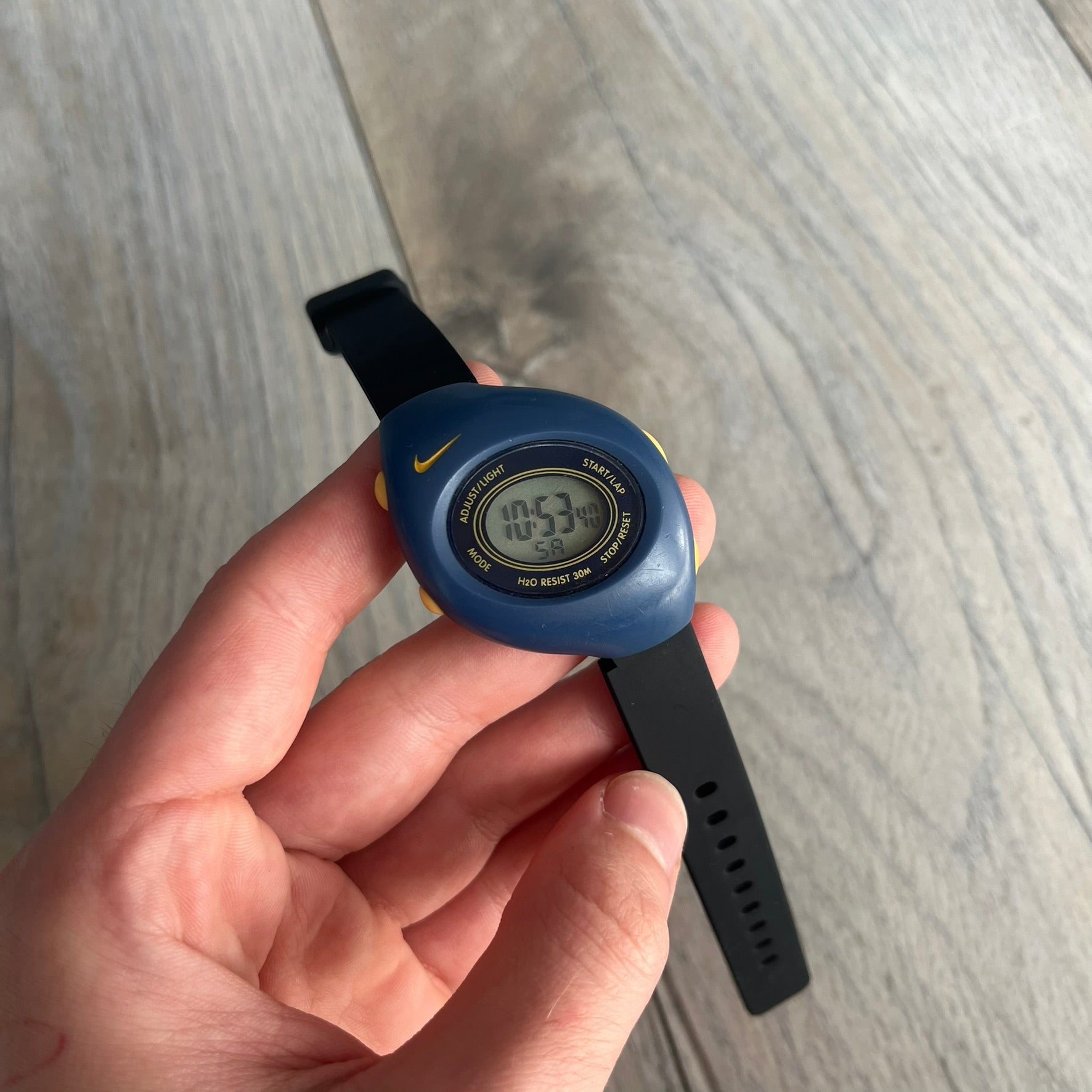 Nike Triax Junior 2000s Blue Digital Watch is a digital timepiece featuring a blue case with a yellow Nike Swoosh on the bezel. The watch includes a backlight with the Bowerman Nike logo, yellow side buttons, and a navy ring around the display with yellow numerals and markings. It has a stainless steel and plastic construction with 30m water resistance. The original strap has been replaced with a black non-Nike strap.