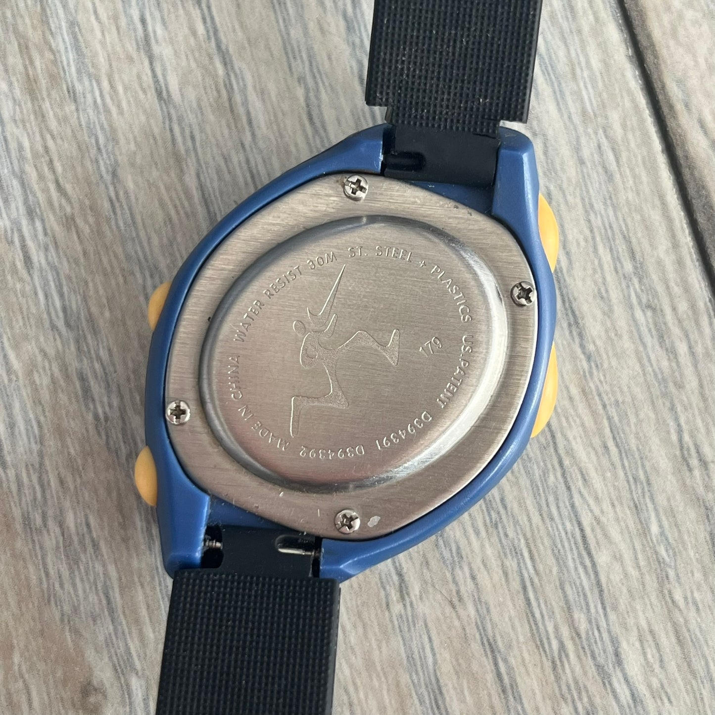 Nike Triax Junior 2000s Blue Digital Watch is a digital timepiece featuring a blue case with a yellow Nike Swoosh on the bezel. The watch includes a backlight with the Bowerman Nike logo, yellow side buttons, and a navy ring around the display with yellow numerals and markings. It has a stainless steel and plastic construction with 30m water resistance. The original strap has been replaced with a black non-Nike strap.