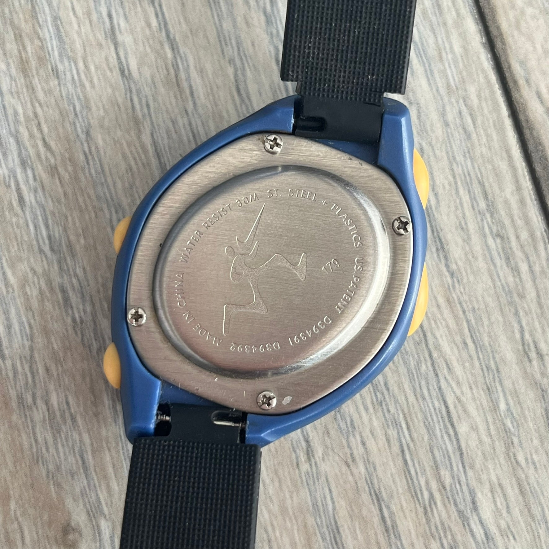 Nike Triax Junior 2000s Blue Digital Watch is a digital timepiece featuring a blue case with a yellow Nike Swoosh on the bezel. The watch includes a backlight with the Bowerman Nike logo, yellow side buttons, and a navy ring around the display with yellow numerals and markings. It has a stainless steel and plastic construction with 30m water resistance. The original strap has been replaced with a black non-Nike strap.