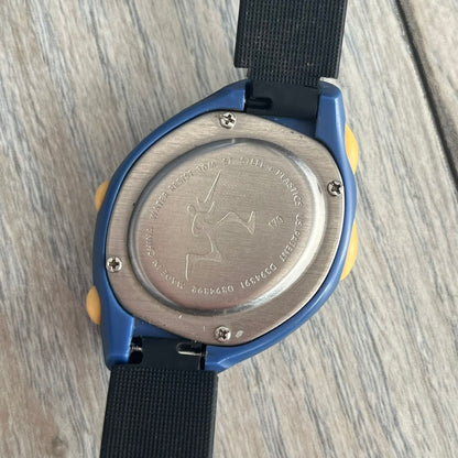 Nike Triax Junior 2000s Blue Digital Watch is a digital timepiece featuring a blue case with a yellow Nike Swoosh on the bezel. The watch includes a backlight with the Bowerman Nike logo, yellow side buttons, and a navy ring around the display with yellow numerals and markings. It has a stainless steel and plastic construction with 30m water resistance. The original strap has been replaced with a black non-Nike strap.