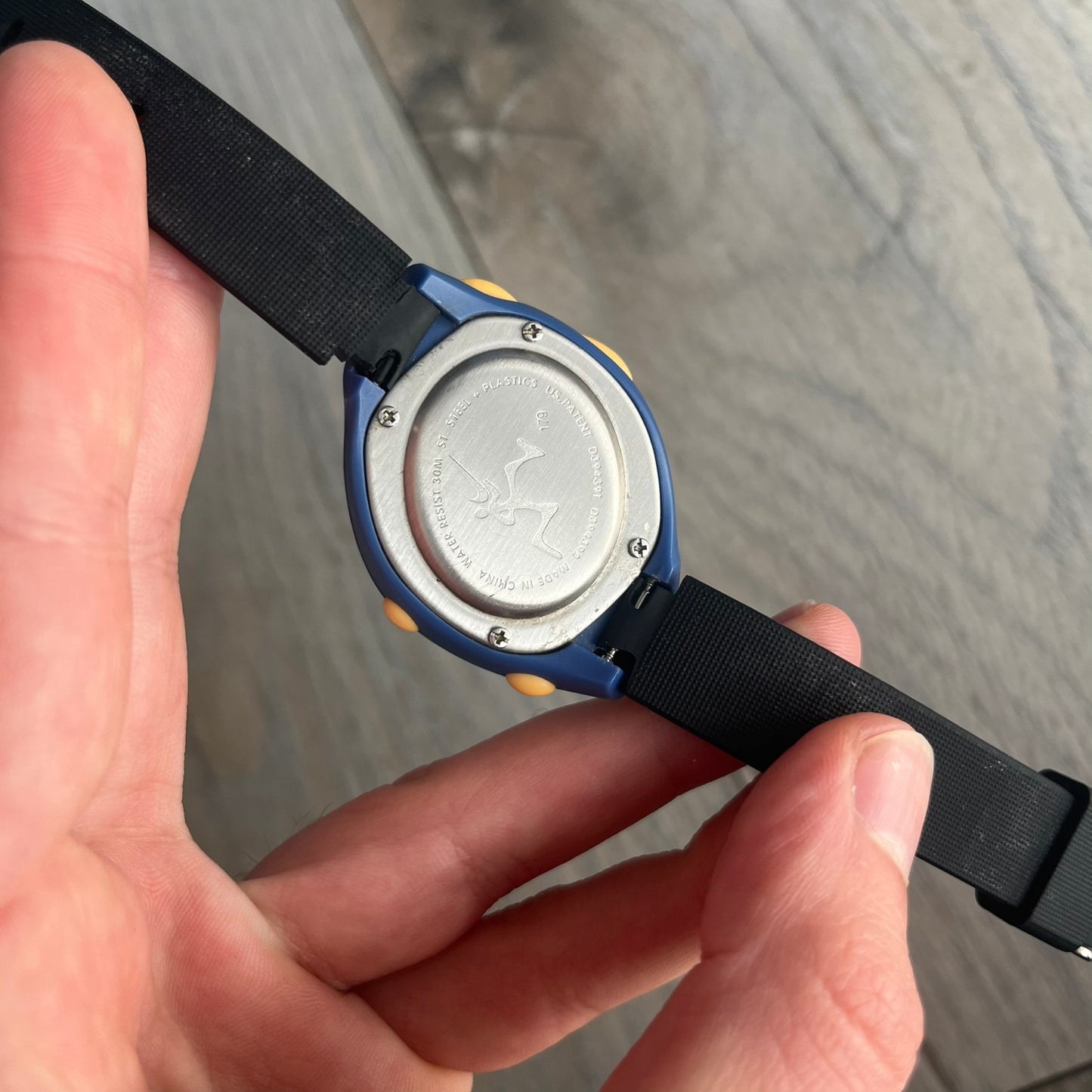 Nike Triax Junior 2000s Blue Digital Watch is a digital timepiece featuring a blue case with a yellow Nike Swoosh on the bezel. The watch includes a backlight with the Bowerman Nike logo, yellow side buttons, and a navy ring around the display with yellow numerals and markings. It has a stainless steel and plastic construction with 30m water resistance. The original strap has been replaced with a black non-Nike strap.