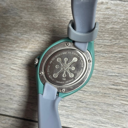 Nike Triax Junior WR0033-401 Fog Blue Analog Watch is a 2000's analog watch featuring a Turquoise case with a small light green Nike logo above the dial. The face is purple and pink with bubbles effect, paired with green hands and pink numerals. It is fitted with gray blue straps. Constructed from stainless steel and plastic materials, the watch is water resistant up to 30 meters. Comes with a tag and rare plastic box.