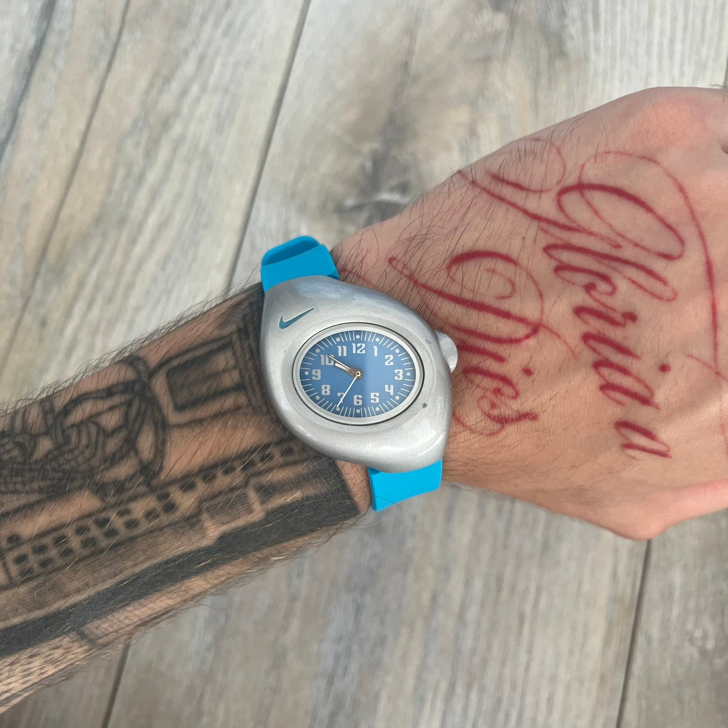 Nike Triax Junior WR0033 Blue Analog Watch is a 2000's analog watch featuring a silver-tone case with a small blue Nike logo above the dial. The light blue face is paired with white hands and white numerals for contrast. It is fitted with blue replacement straps that are not original Nike straps. Constructed from stainless steel and plastic materials, the watch is water resistant up to 50 meters and includes US patent markings.