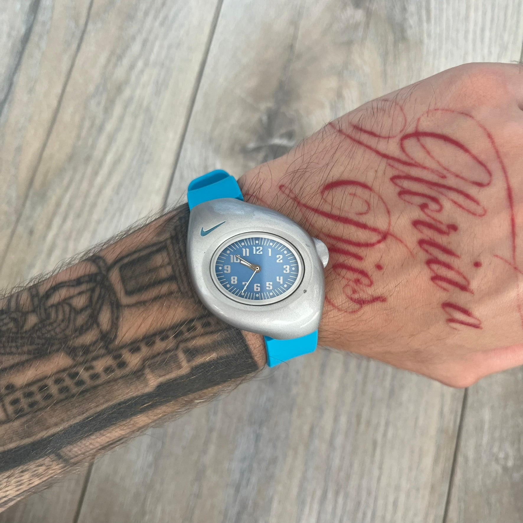 Nike Triax Junior WR0033 Blue Analog Watch is a 2000's analog watch featuring a silver-tone case with a small blue Nike logo above the dial. The light blue face is paired with white hands and white numerals for contrast. It is fitted with blue replacement straps that are not original Nike straps. Constructed from stainless steel and plastic materials, the watch is water resistant up to 50 meters and includes US patent markings.