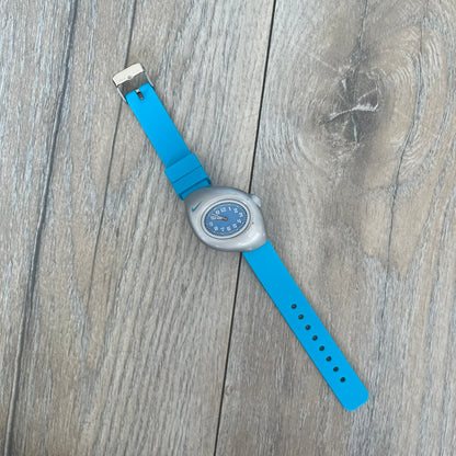 Nike Triax Junior WR0033 Blue Analog Watch is a 2000's analog watch featuring a silver-tone case with a small blue Nike logo above the dial. The light blue face is paired with white hands and white numerals for contrast. It is fitted with blue replacement straps that are not original Nike straps. Constructed from stainless steel and plastic materials, the watch is water resistant up to 50 meters and includes US patent markings.
