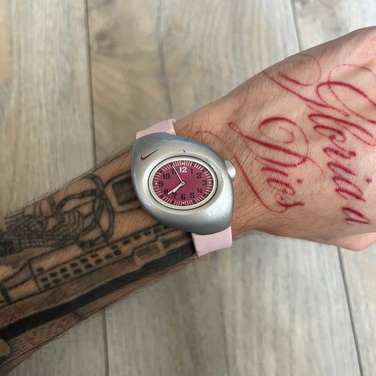 Nike Triax Junior WR0033 Pink Analog Watch is a 2000s analog watch featuring a silver-tone case with a small dark pink Nike logo above the dial. The dark pink face is paired with white hands and white-and-black numerals for contrast. It is fitted with light pink replacement straps that are not original Nike straps. Constructed from stainless steel and plastic materials, the watch is water resistant up to 50 meters and includes US patent markings.
