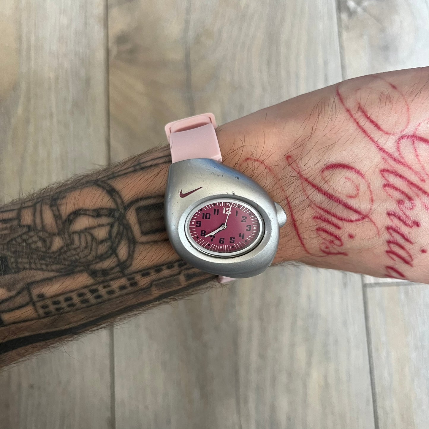 Nike Triax Junior WR0033 Pink Analog Watch is a 2000s analog watch featuring a silver-tone case with a small dark pink Nike logo above the dial. The dark pink face is paired with white hands and white-and-black numerals for contrast. It is fitted with light pink replacement straps that are not original Nike straps. Constructed from stainless steel and plastic materials, the watch is water resistant up to 50 meters and includes US patent markings.