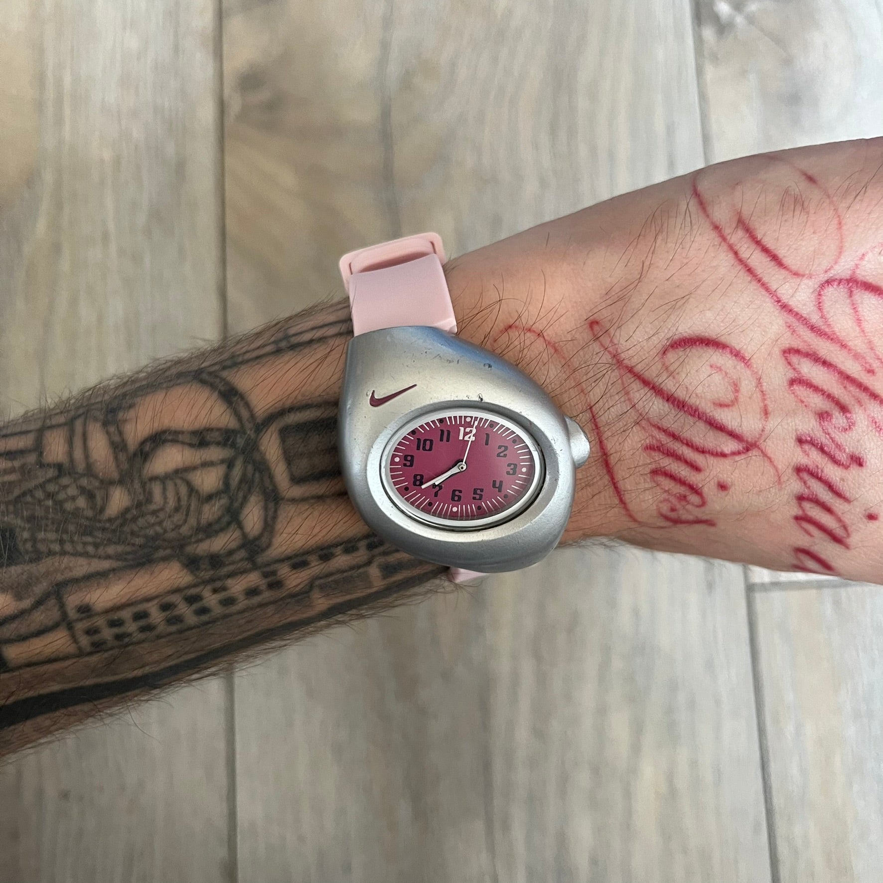 Nike Triax Junior WR0033 Pink Analog Watch is a 2000s analog watch featuring a silver-tone case with a small dark pink Nike logo above the dial. The dark pink face is paired with white hands and white-and-black numerals for contrast. It is fitted with light pink replacement straps that are not original Nike straps. Constructed from stainless steel and plastic materials, the watch is water resistant up to 50 meters and includes US patent markings.