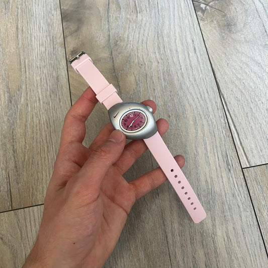 Nike Triax Junior WR0033 Pink Analog Watch is a 2000s analog watch featuring a silver-tone case with a small dark pink Nike logo above the dial. The dark pink face is paired with white hands and white-and-black numerals for contrast. It is fitted with light pink replacement straps that are not original Nike straps. Constructed from stainless steel and plastic materials, the watch is water resistant up to 50 meters and includes US patent markings.