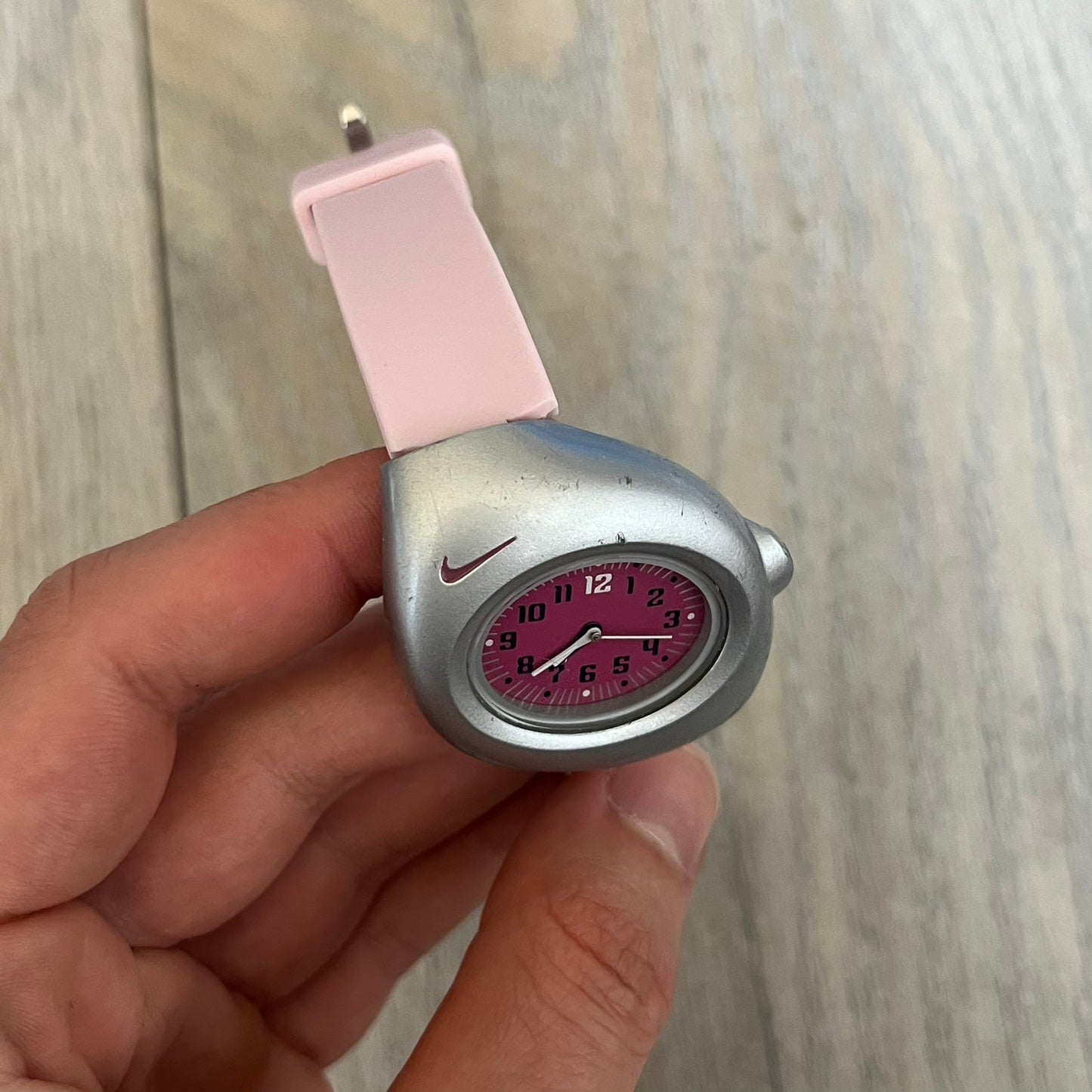 Nike Triax Junior WR0033 Pink Analog Watch is a 2000s analog watch featuring a silver-tone case with a small dark pink Nike logo above the dial. The dark pink face is paired with white hands and white-and-black numerals for contrast. It is fitted with light pink replacement straps that are not original Nike straps. Constructed from stainless steel and plastic materials, the watch is water resistant up to 50 meters and includes US patent markings.