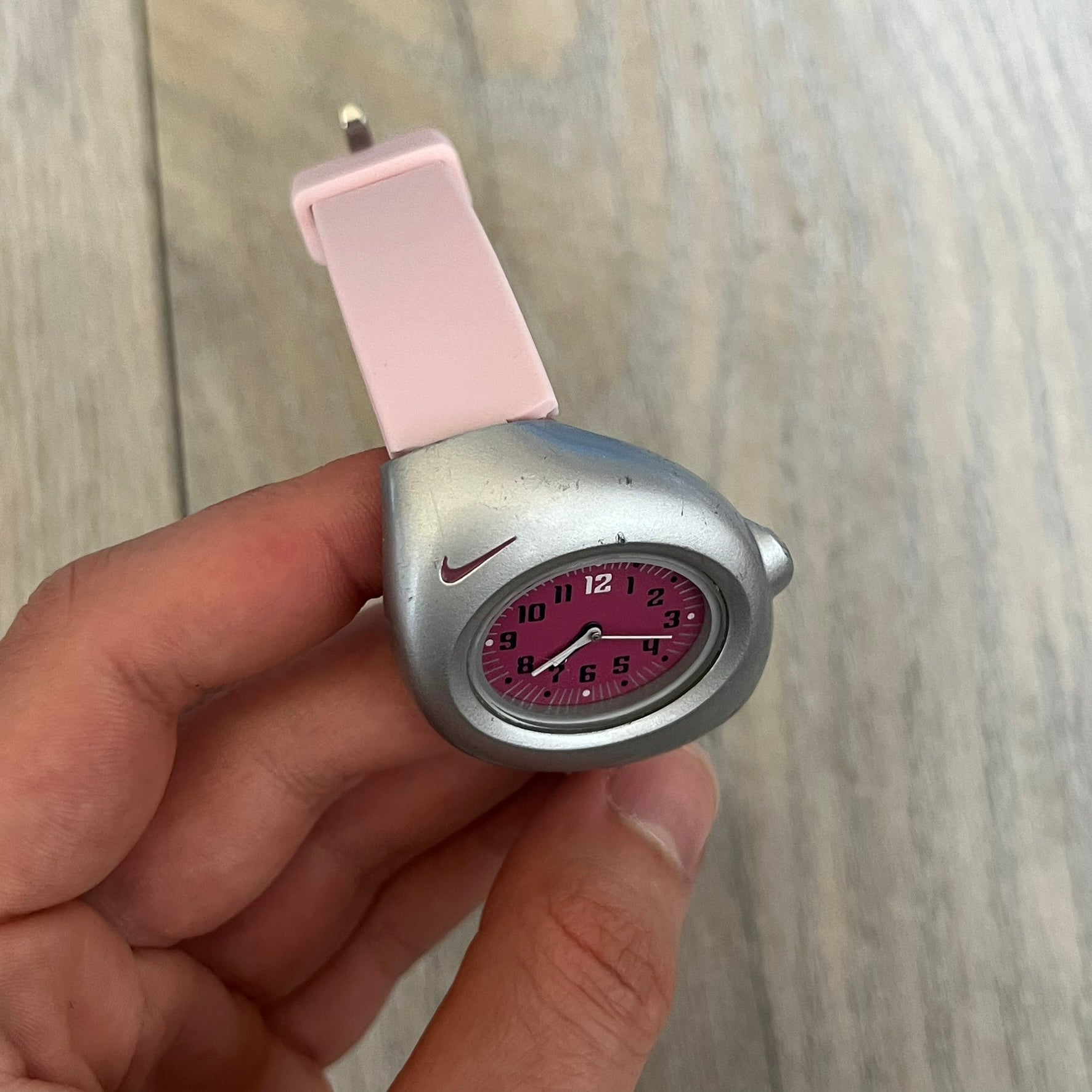 Nike Triax Junior WR0033 Pink Analog Watch is a 2000s analog watch featuring a silver-tone case with a small dark pink Nike logo above the dial. The dark pink face is paired with white hands and white-and-black numerals for contrast. It is fitted with light pink replacement straps that are not original Nike straps. Constructed from stainless steel and plastic materials, the watch is water resistant up to 50 meters and includes US patent markings.