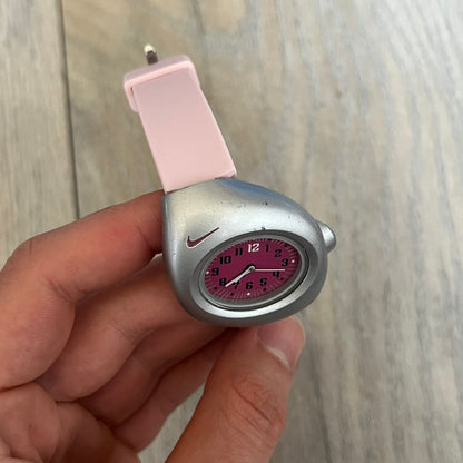 Nike Triax Junior WR0033 Pink Analog Watch is a 2000s analog watch featuring a silver-tone case with a small dark pink Nike logo above the dial. The dark pink face is paired with white hands and white-and-black numerals for contrast. It is fitted with light pink replacement straps that are not original Nike straps. Constructed from stainless steel and plastic materials, the watch is water resistant up to 50 meters and includes US patent markings.
