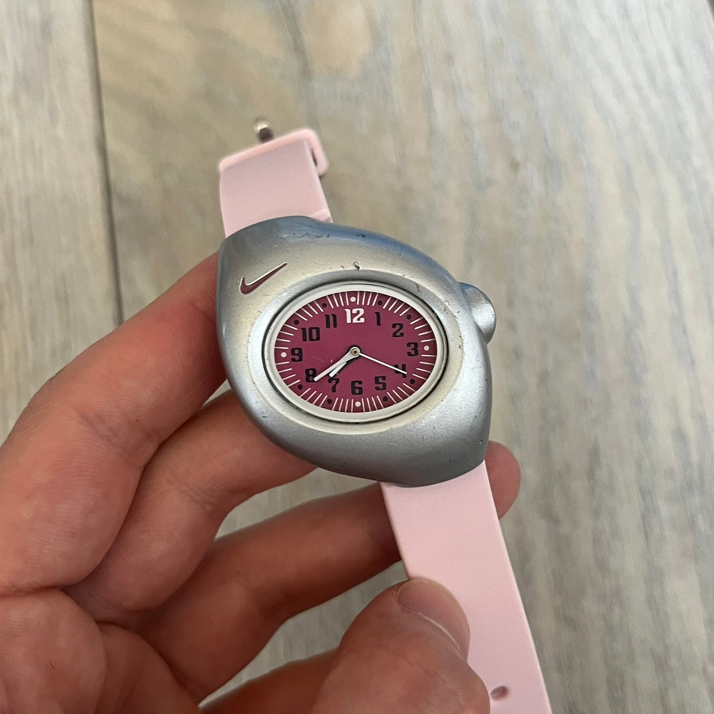 Nike Triax Junior WR0033 Pink Analog Watch is a 2000s analog watch featuring a silver-tone case with a small dark pink Nike logo above the dial. The dark pink face is paired with white hands and white-and-black numerals for contrast. It is fitted with light pink replacement straps that are not original Nike straps. Constructed from stainless steel and plastic materials, the watch is water resistant up to 50 meters and includes US patent markings.