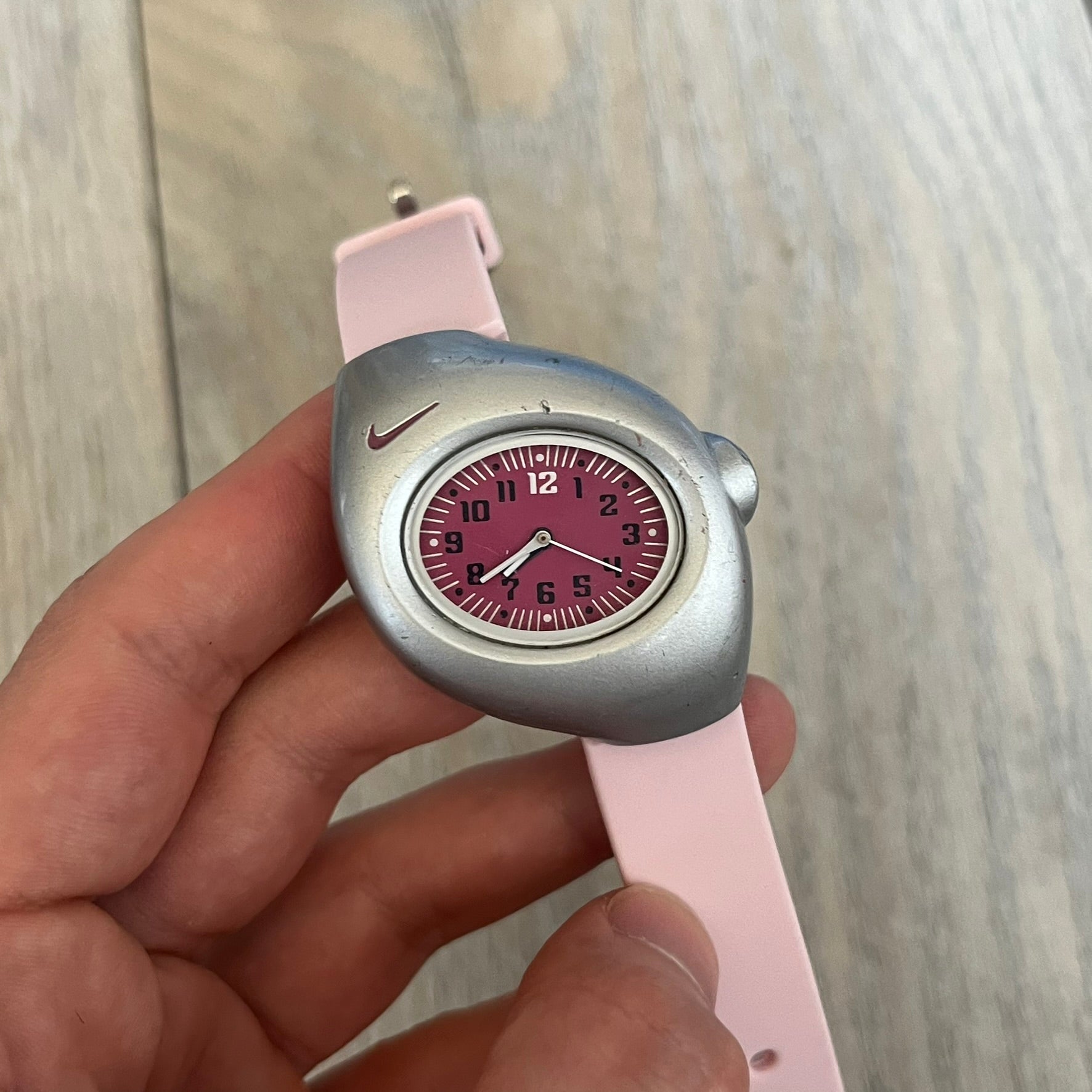 Nike Triax Junior WR0033 Pink Analog Watch is a 2000s analog watch featuring a silver-tone case with a small dark pink Nike logo above the dial. The dark pink face is paired with white hands and white-and-black numerals for contrast. It is fitted with light pink replacement straps that are not original Nike straps. Constructed from stainless steel and plastic materials, the watch is water resistant up to 50 meters and includes US patent markings.