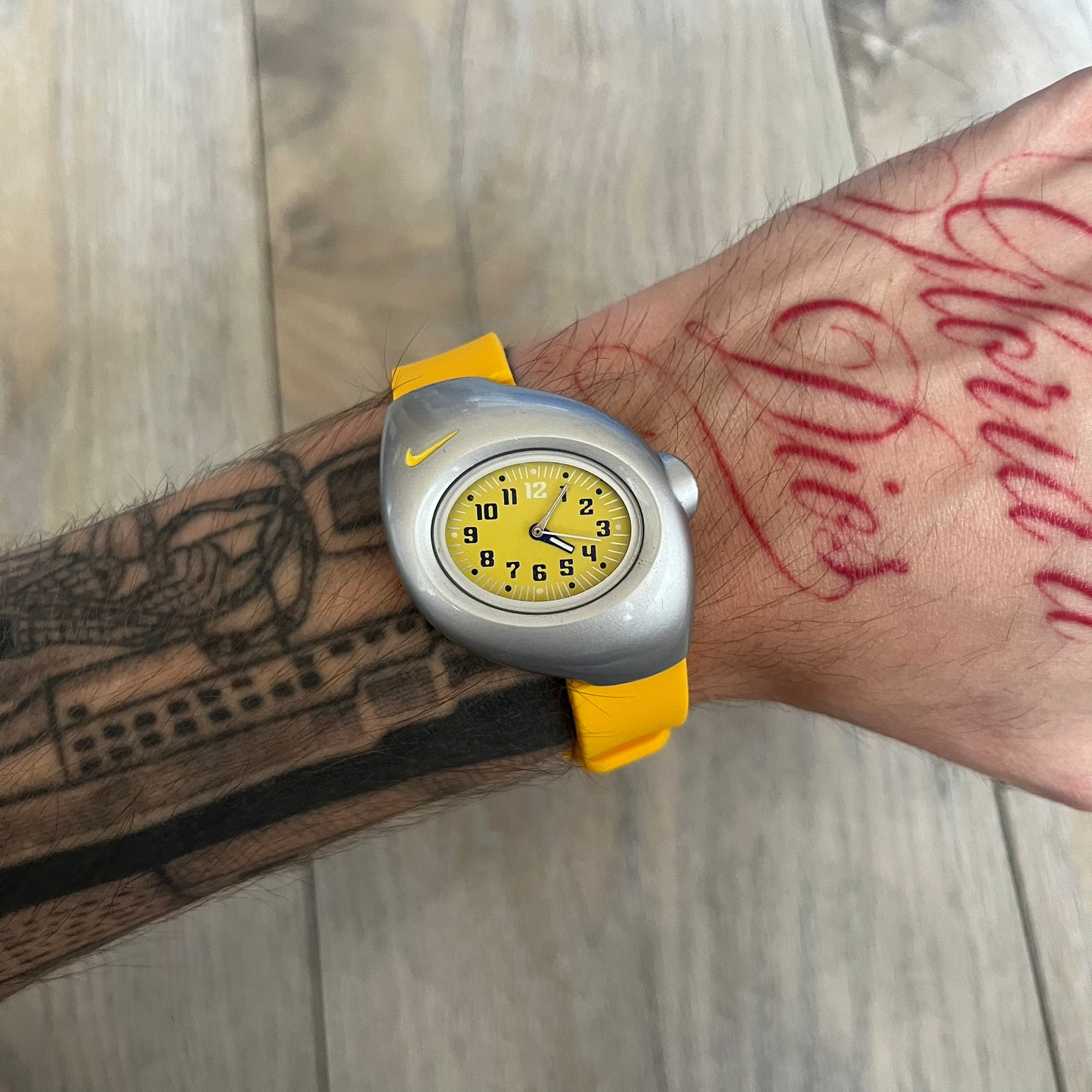 Nike Triax Junior WR0033 Yellow Analog Watch is a 2000s analog watch featuring a silver-tone case with a small yellow Nike logo above the dial. The yellow face is paired with white hands and white-and-black numerals for contrast. It is fitted with yellow replacement straps that are not original Nike straps. Constructed from stainless steel and plastic materials, the watch is water resistant up to 50 meters and includes US patent markings.