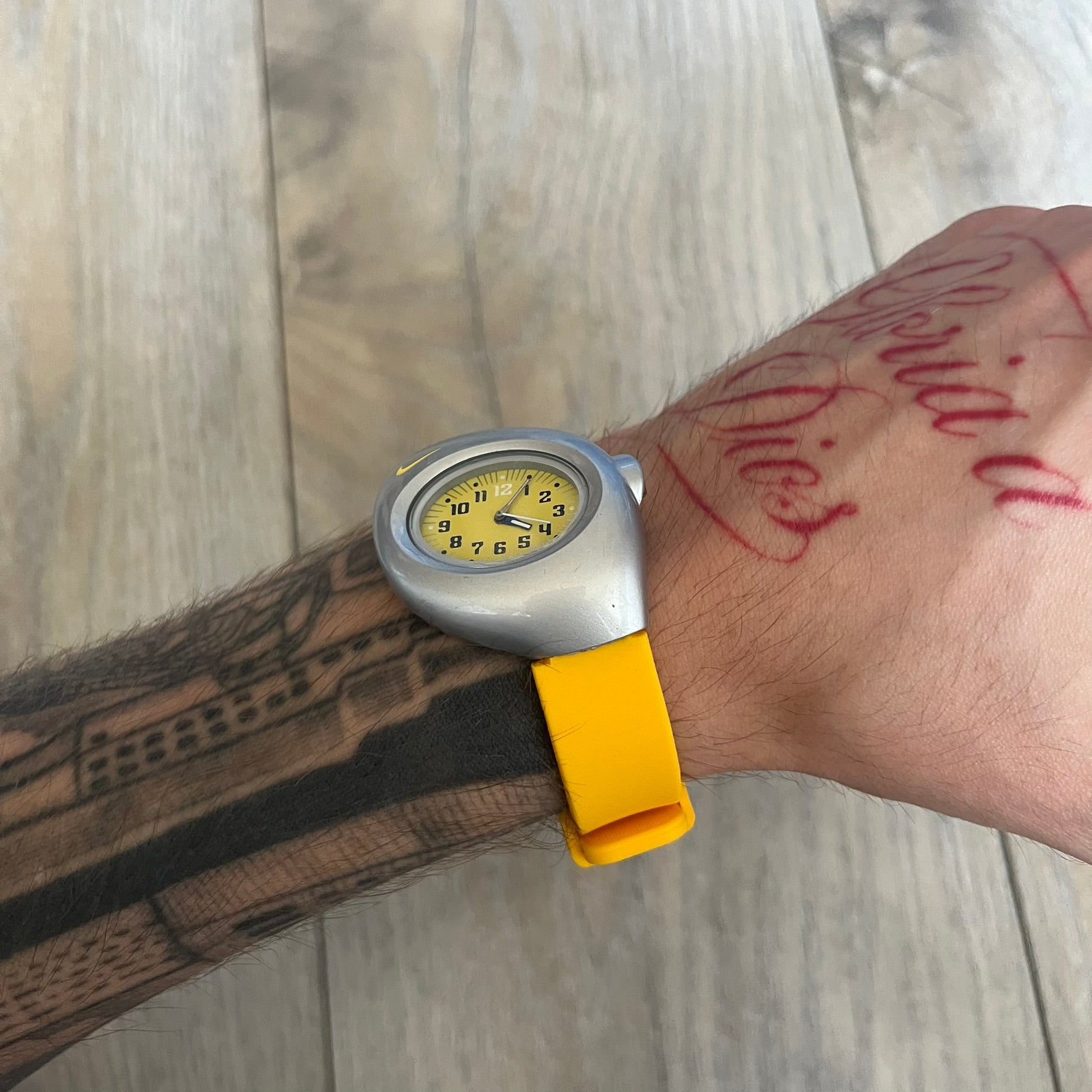 Nike Triax Junior WR0033 Yellow Analog Watch is a 2000s analog watch featuring a silver-tone case with a small yellow Nike logo above the dial. The yellow face is paired with white hands and white-and-black numerals for contrast. It is fitted with yellow replacement straps that are not original Nike straps. Constructed from stainless steel and plastic materials, the watch is water resistant up to 50 meters and includes US patent markings.